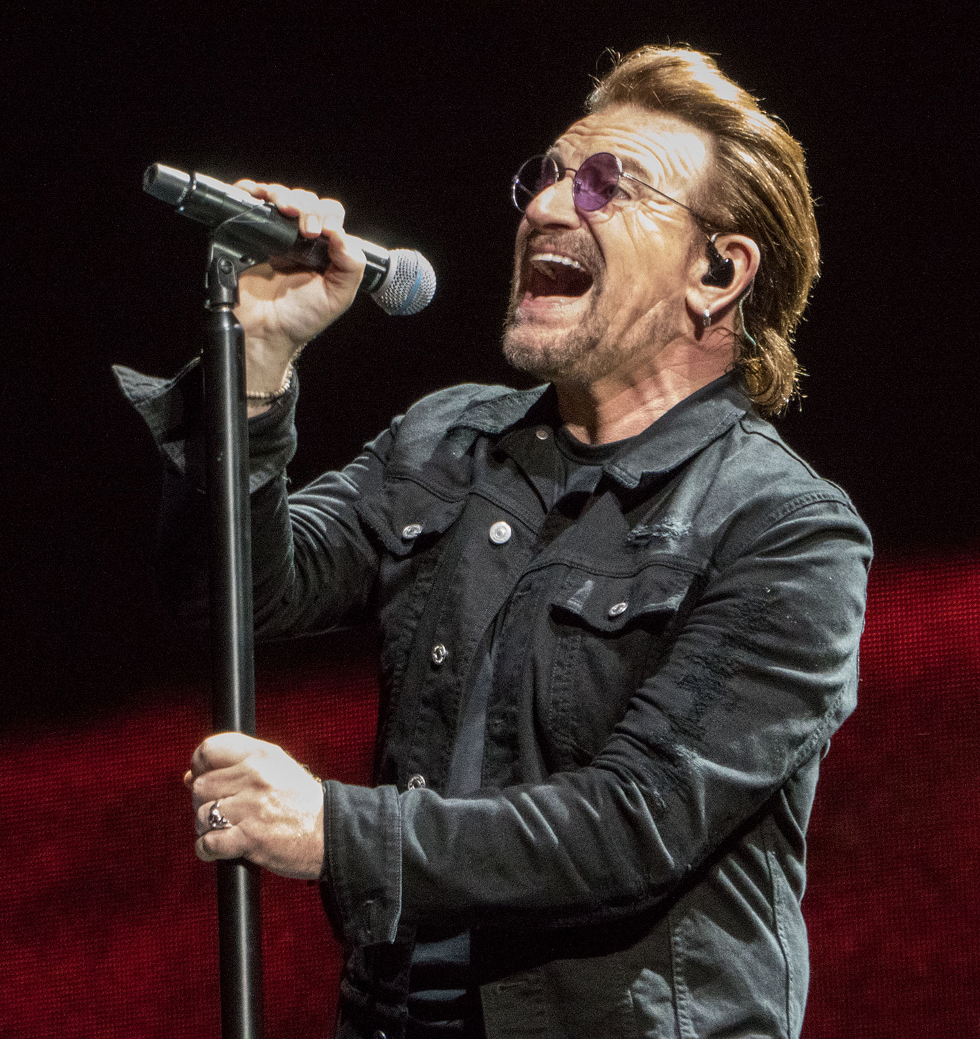 Bono sings into a microphone on a stand with his mouth wide open, wearing round tinted glasses and a dark denim jacket, against a deep red backdrop.