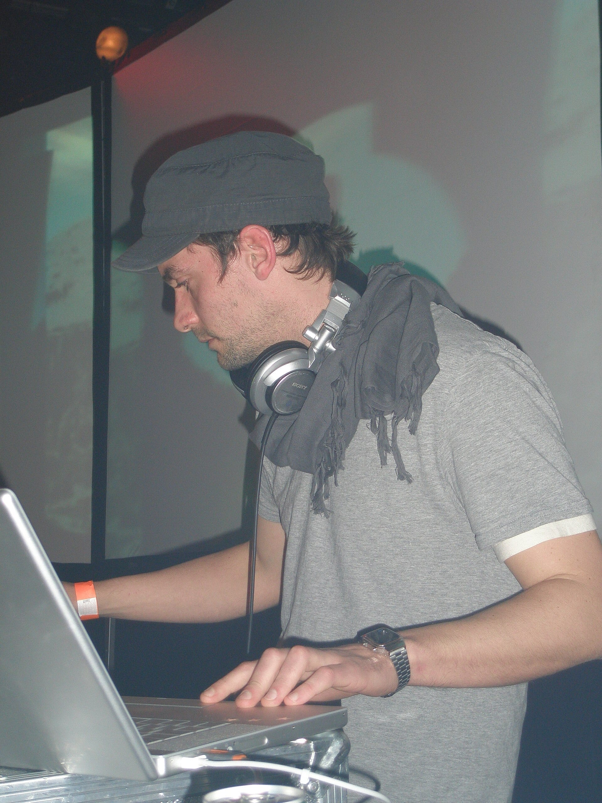 Simon Green, who performs as Bonobo, wearing a flat cap and headphones while DJing at a club.