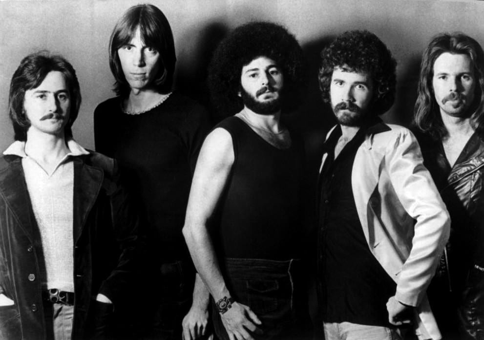A black and white studio portrait of the five members of Boston standing shoulder to shoulder in front of a plain wall, in denim, leather and open-collared shirts.