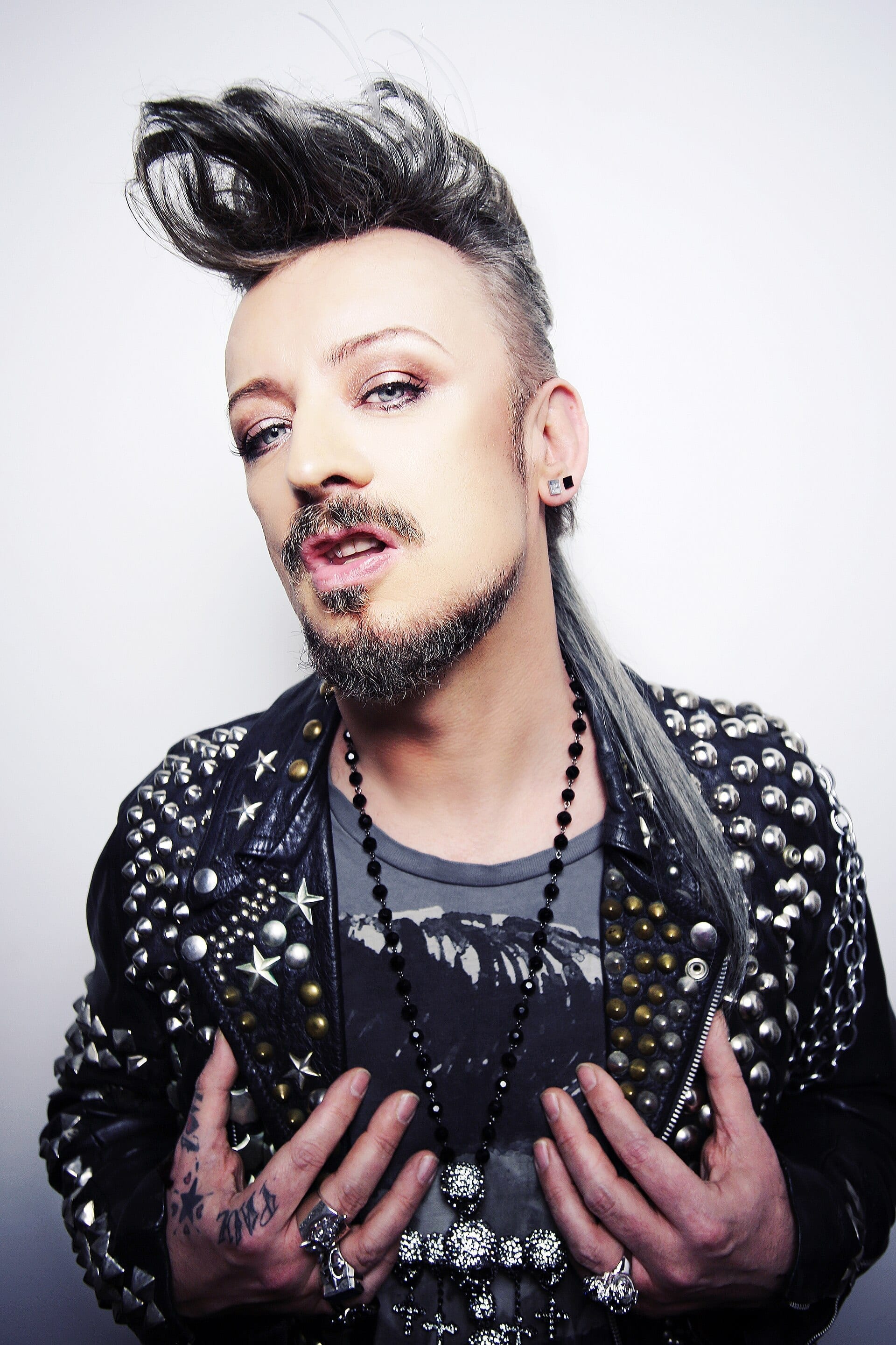 A studio portrait of Boy George with a swept-up black and gray mohawk, dramatic eye makeup, and a studded leather jacket over a graphic T-shirt, hands clasped at his chest.