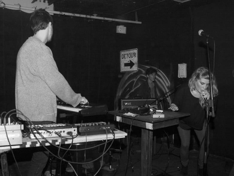 A black and white shot of a small club stage: a man plays a synth rig at left while a woman doubles over into a microphone at right, a road-sign arrow on the wall between them.