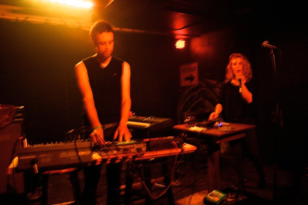 Two performers on a small dark stage under red light, one standing over a synthesiser rig and the other singing into a handheld microphone beside a drum machine on a table