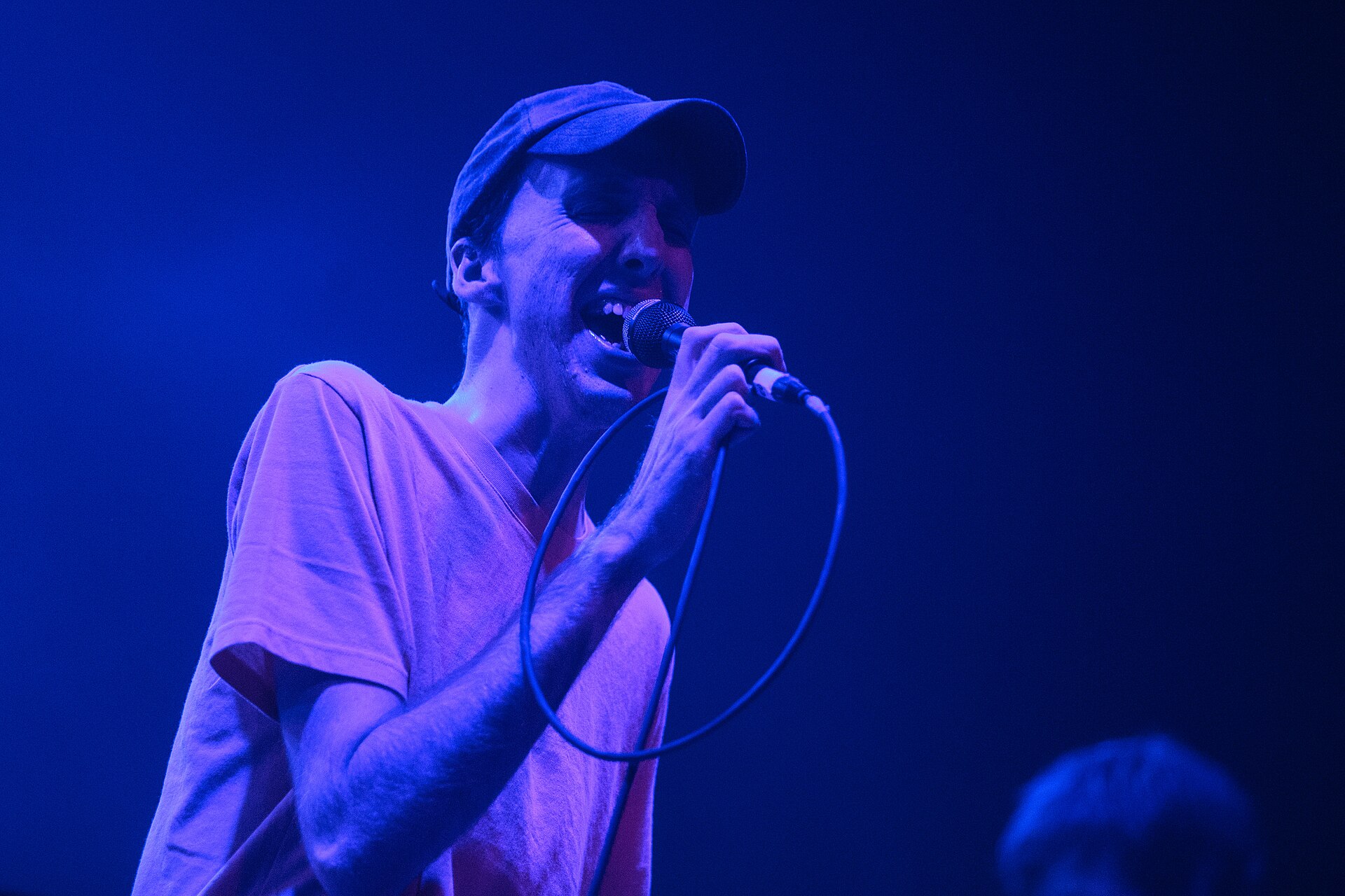 Bradford Cox singing into a handheld microphone under deep blue stage light, in a soft cap and a loose pale t-shirt, the mic cable looped around his arm