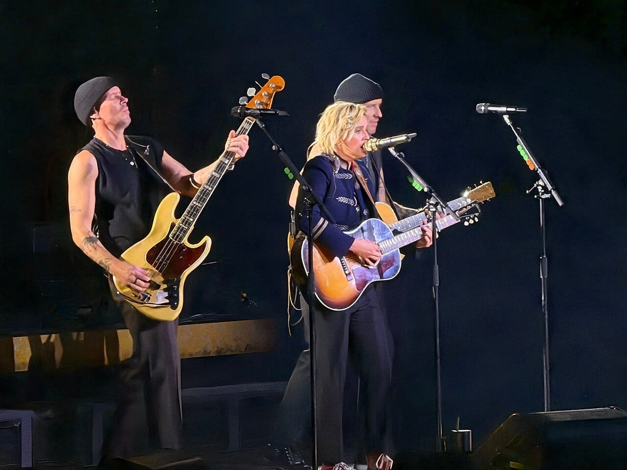 Brandi Carlile sings at a microphone while playing acoustic guitar, with two bandmates beside her on a dark stage.