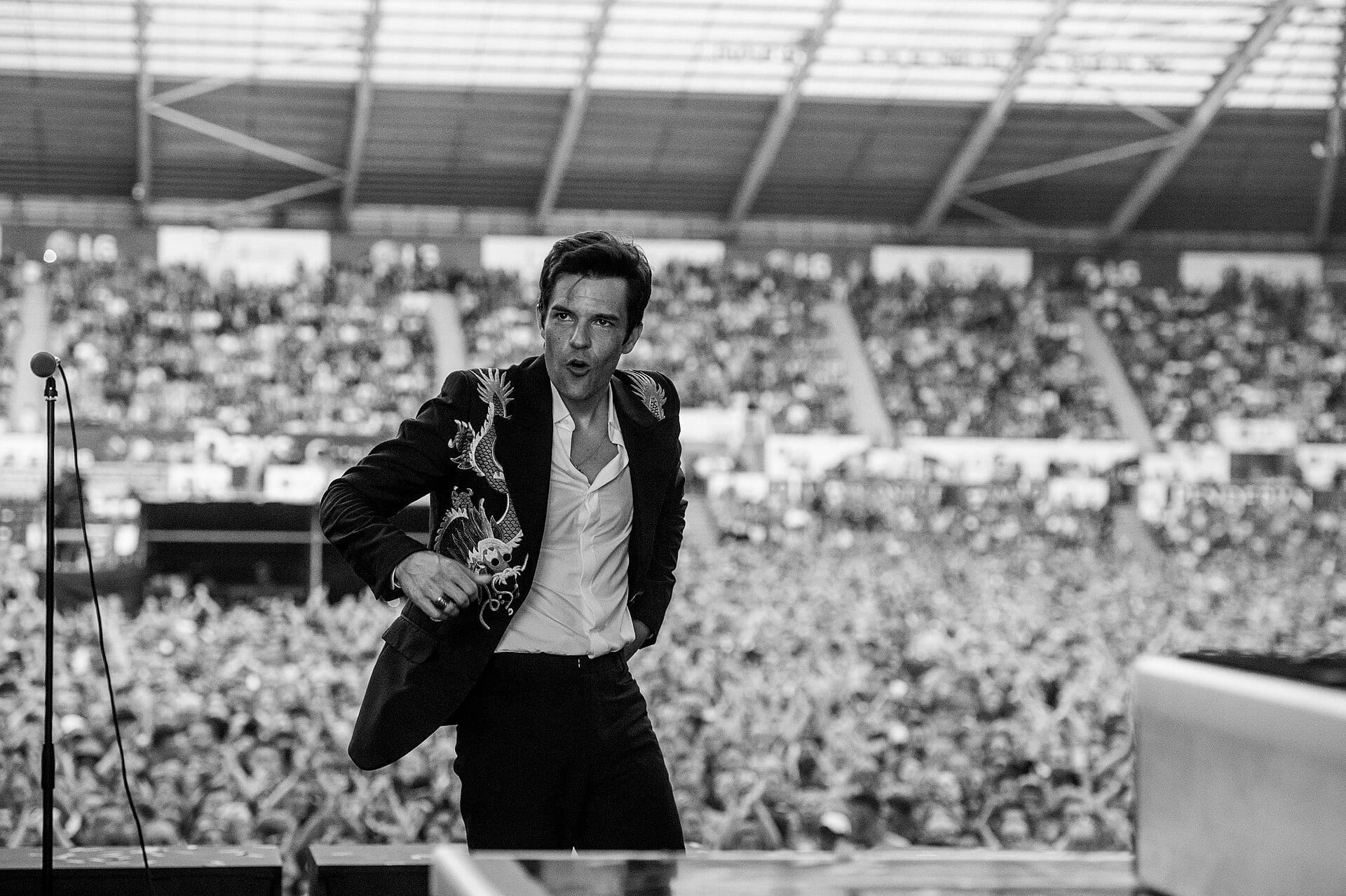 A black-and-white photo of Brandon Flowers performing on an outdoor stage in a dragon-embroidered blazer and open white shirt, with a packed stadium crowd behind him.