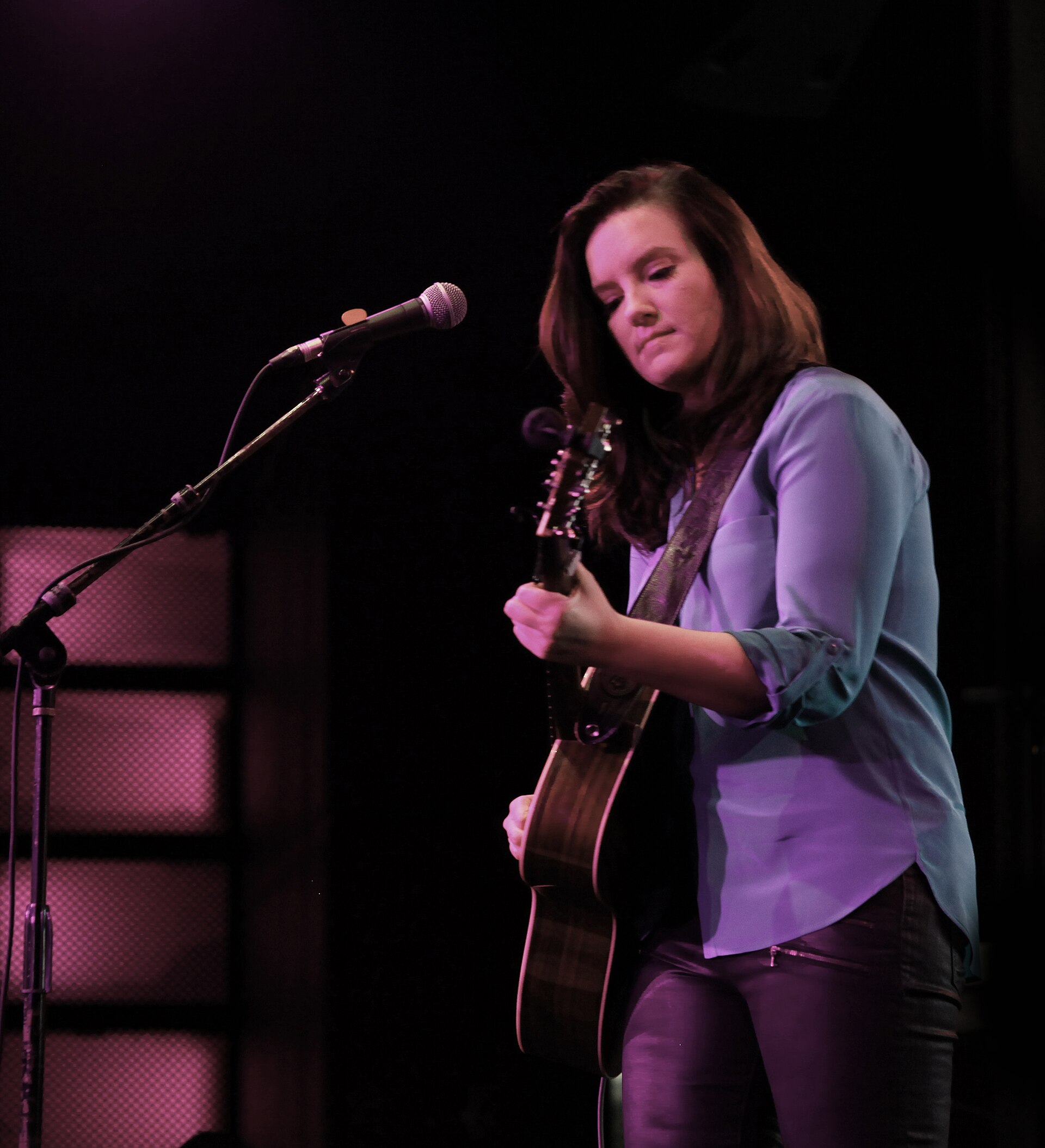 Brandy Clark plays an acoustic guitar on a dark club stage, head down and eyes lowered, a microphone on a stand to her right under purple light.