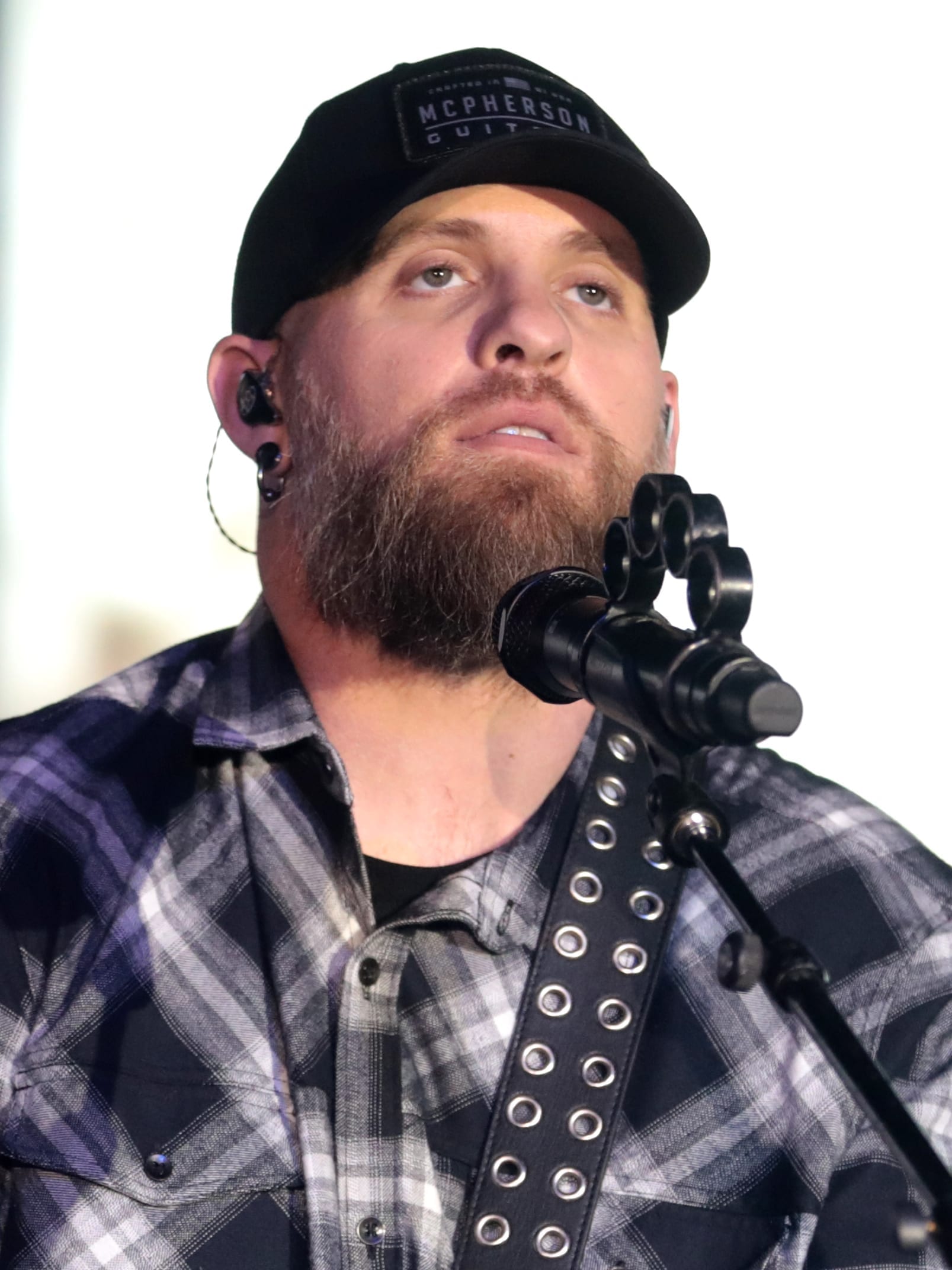 Brantley Gilbert performs at a microphone, wearing a black cap and plaid shirt, with a full beard.
