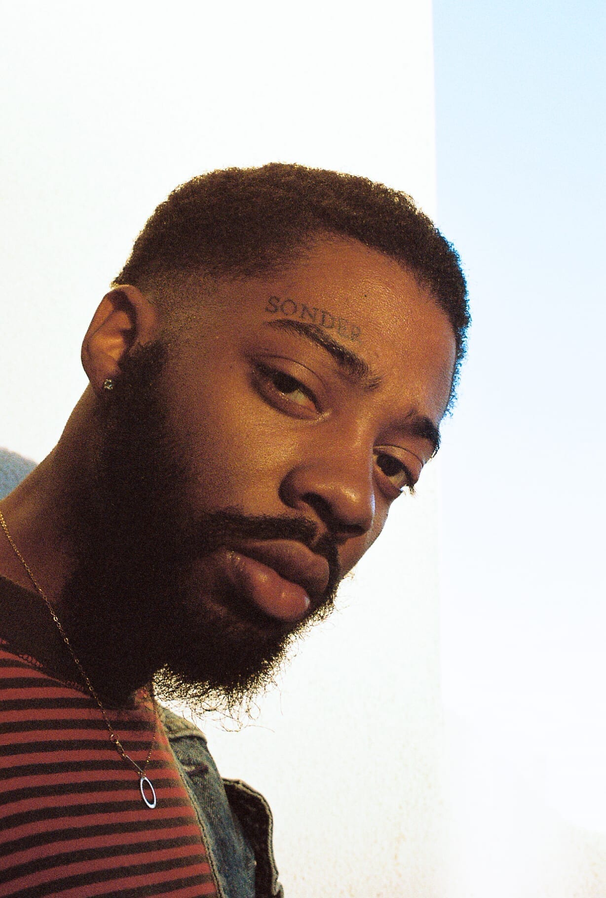 Close-up of Brent Faiyaz looking downward, wearing a striped shirt and a gold necklace, with the word Sonder tattooed above his eyebrow.