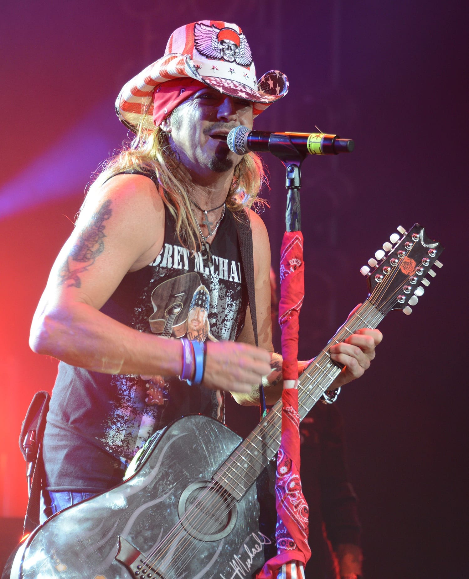 Bret Michaels sings into a microphone while playing an acoustic guitar, wearing a stars-and-stripes cowboy hat over a red bandana.