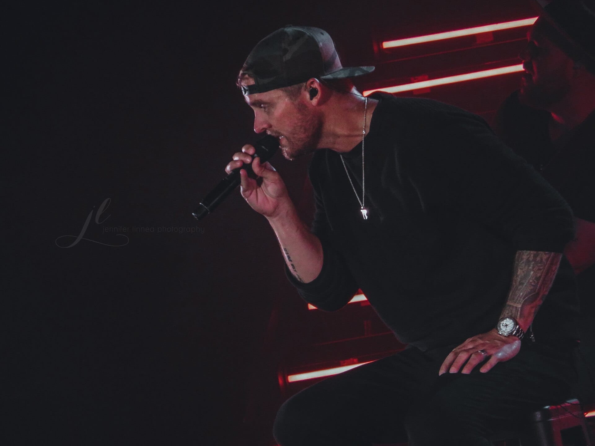 Brett Young sings into a microphone in a backwards cap and dark shirt, an acoustic guitar strap over his shoulder.