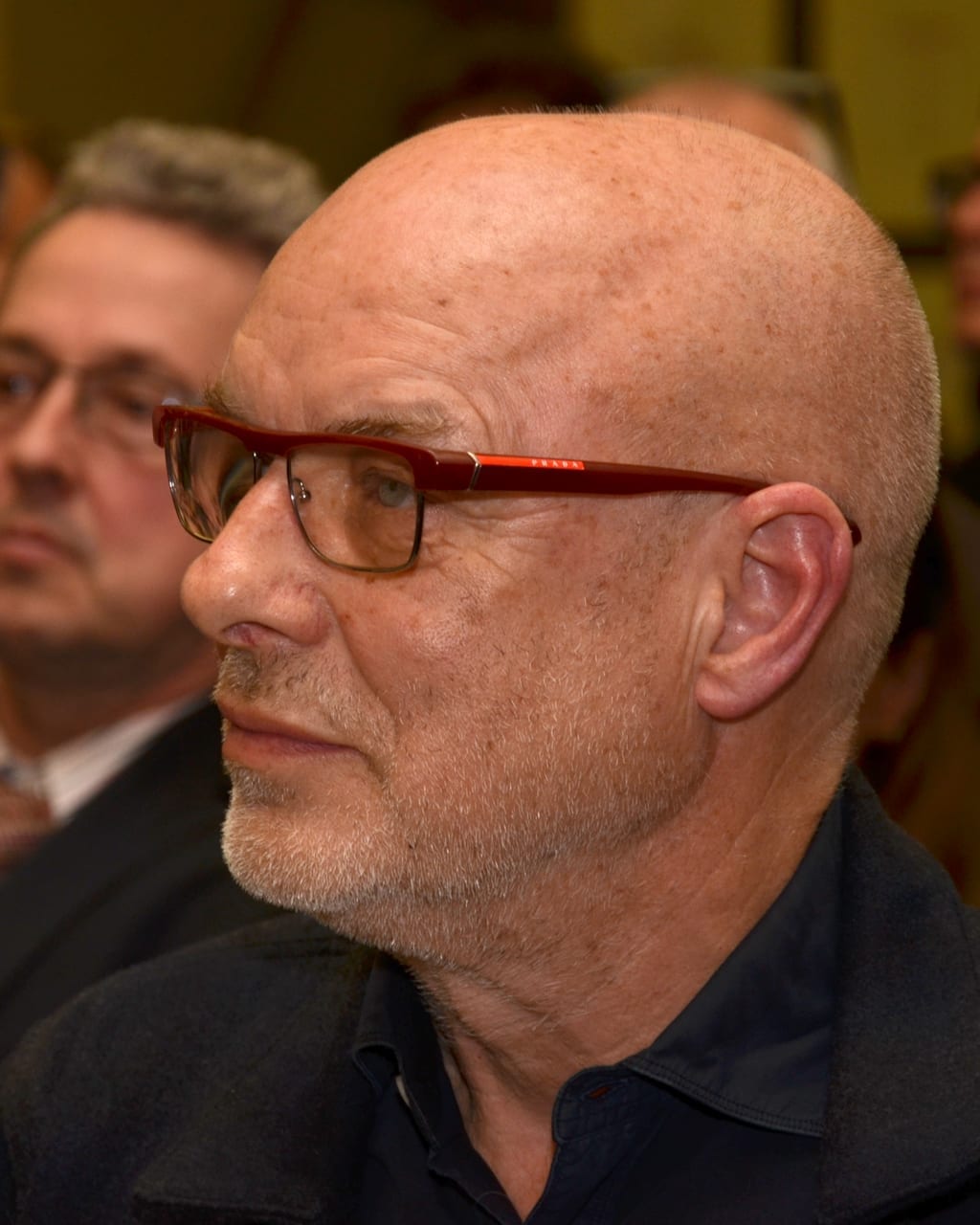 Brian Eno, bald with red-framed glasses and a gray beard, is seen in profile in a dark shirt.