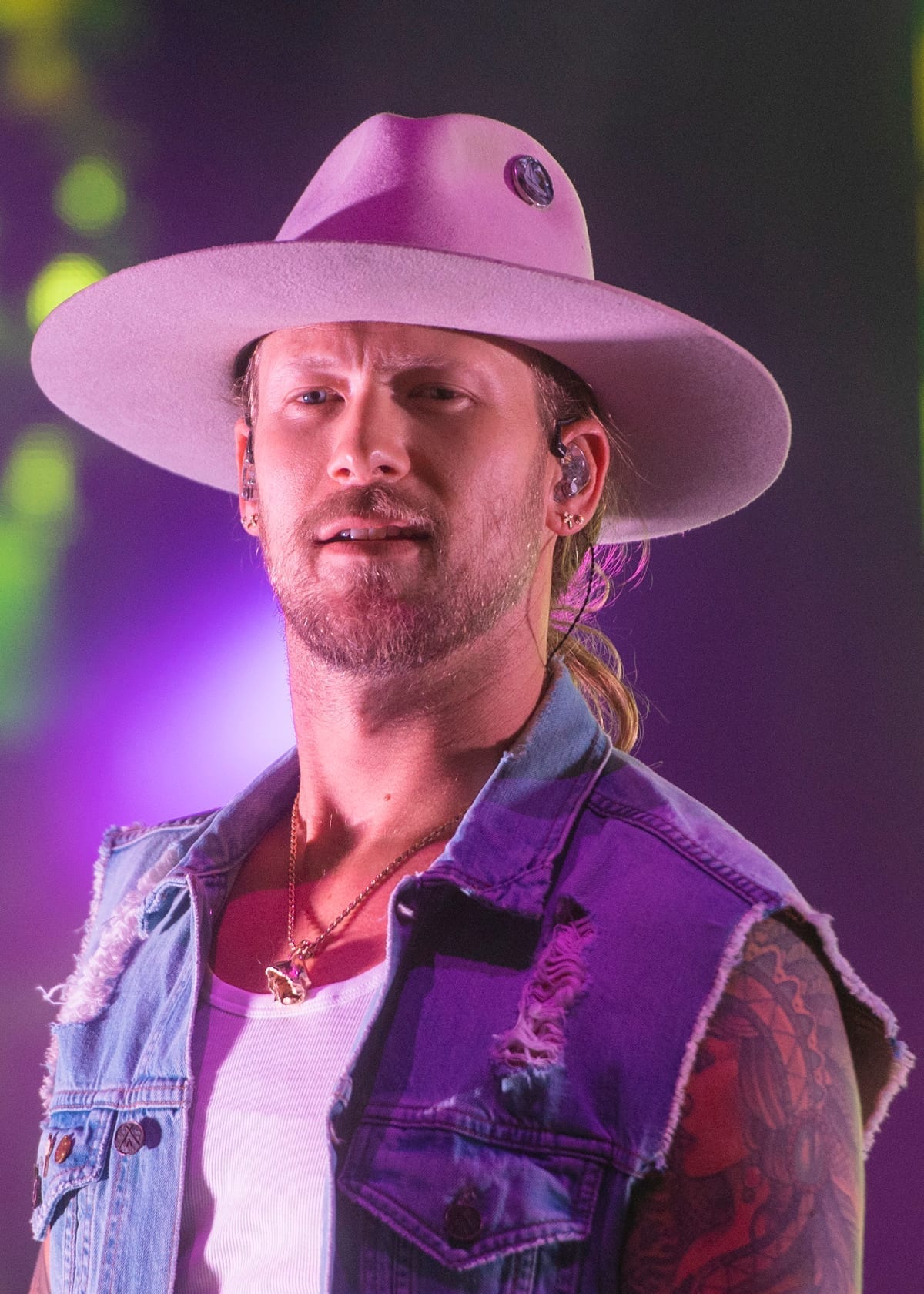 Brian Kelley, wearing a wide-brimmed hat and a denim vest over tattooed arms, looks out during a live performance.