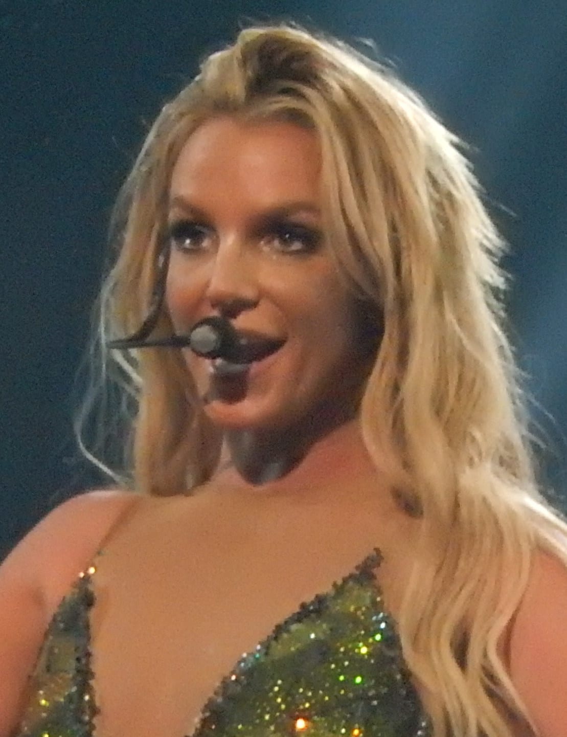 Britney Spears, close up, sings into a microphone in a glittery green top, her blonde hair falling loose around her shoulders.