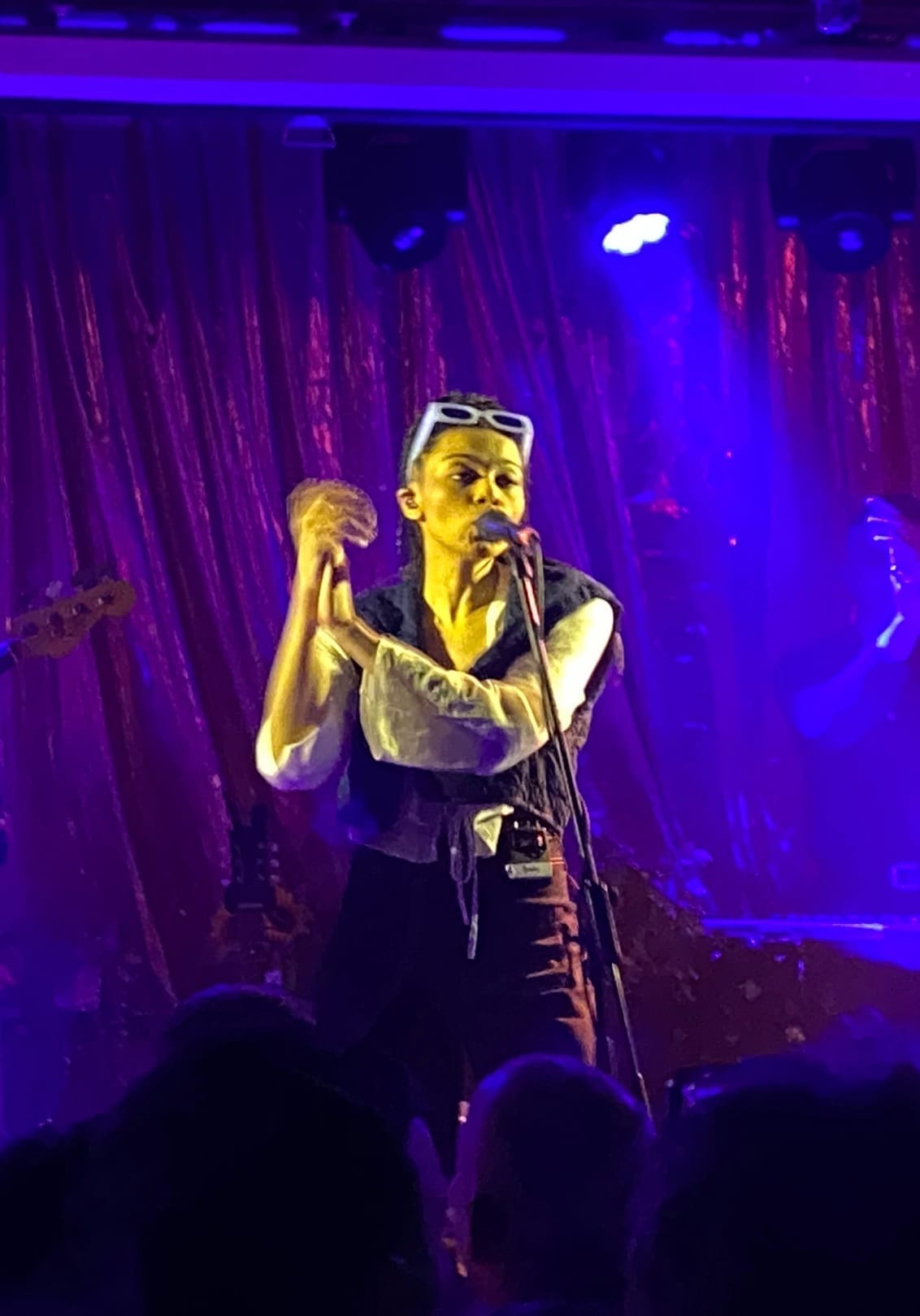 Brooke Combe sings into a microphone under purple stage light, sunglasses pushed up into her hair, hands raised near her chest.