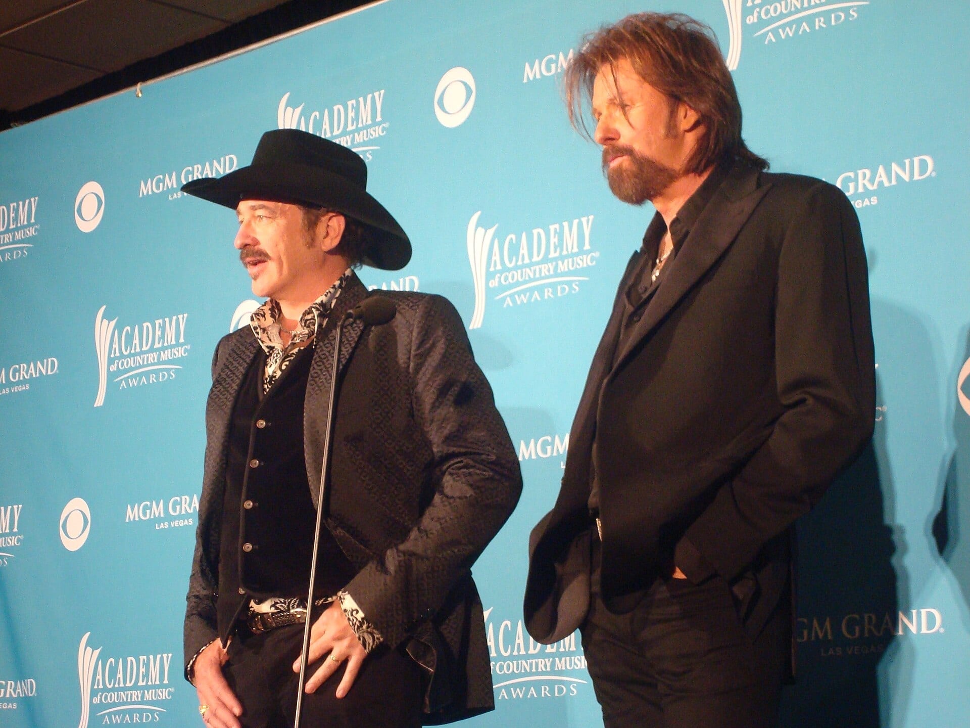 Kix Brooks, in a black cowboy hat, and Ronnie Dunn, bearded in a black suit, stand together at a step-and-repeat backdrop for the Academy of Country Music Awards.