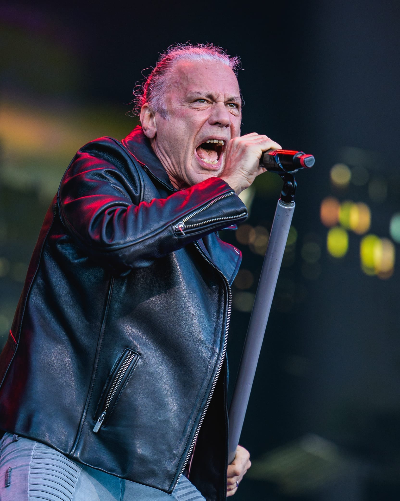 Bruce Dickinson leans hard into a microphone on its stand, mouth wide open mid-scream, in a black leather jacket under red light.