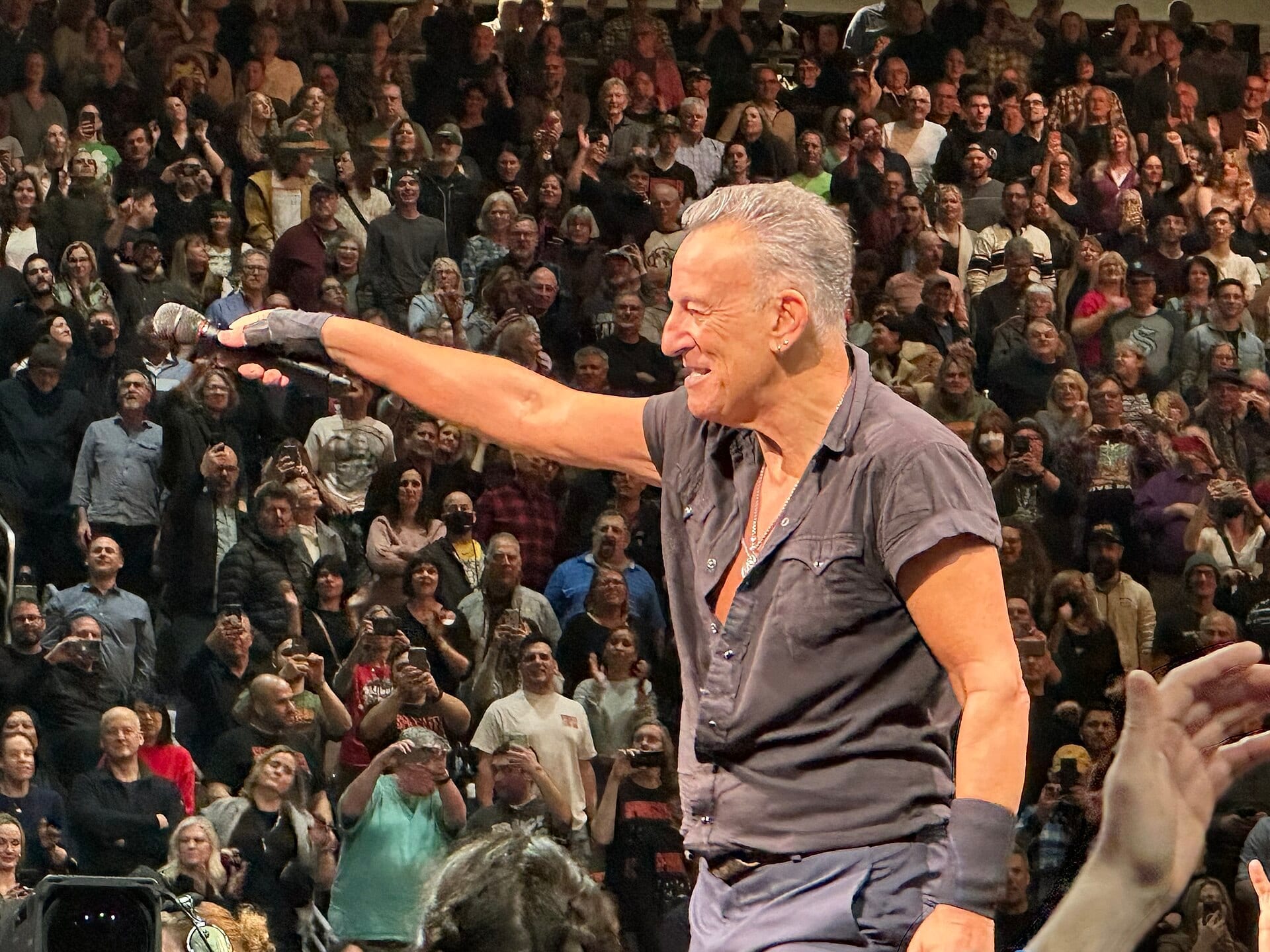Bruce Springsteen grins and holds a microphone out toward the crowd, sleeves rolled up, a packed arena tier filling the frame behind him.