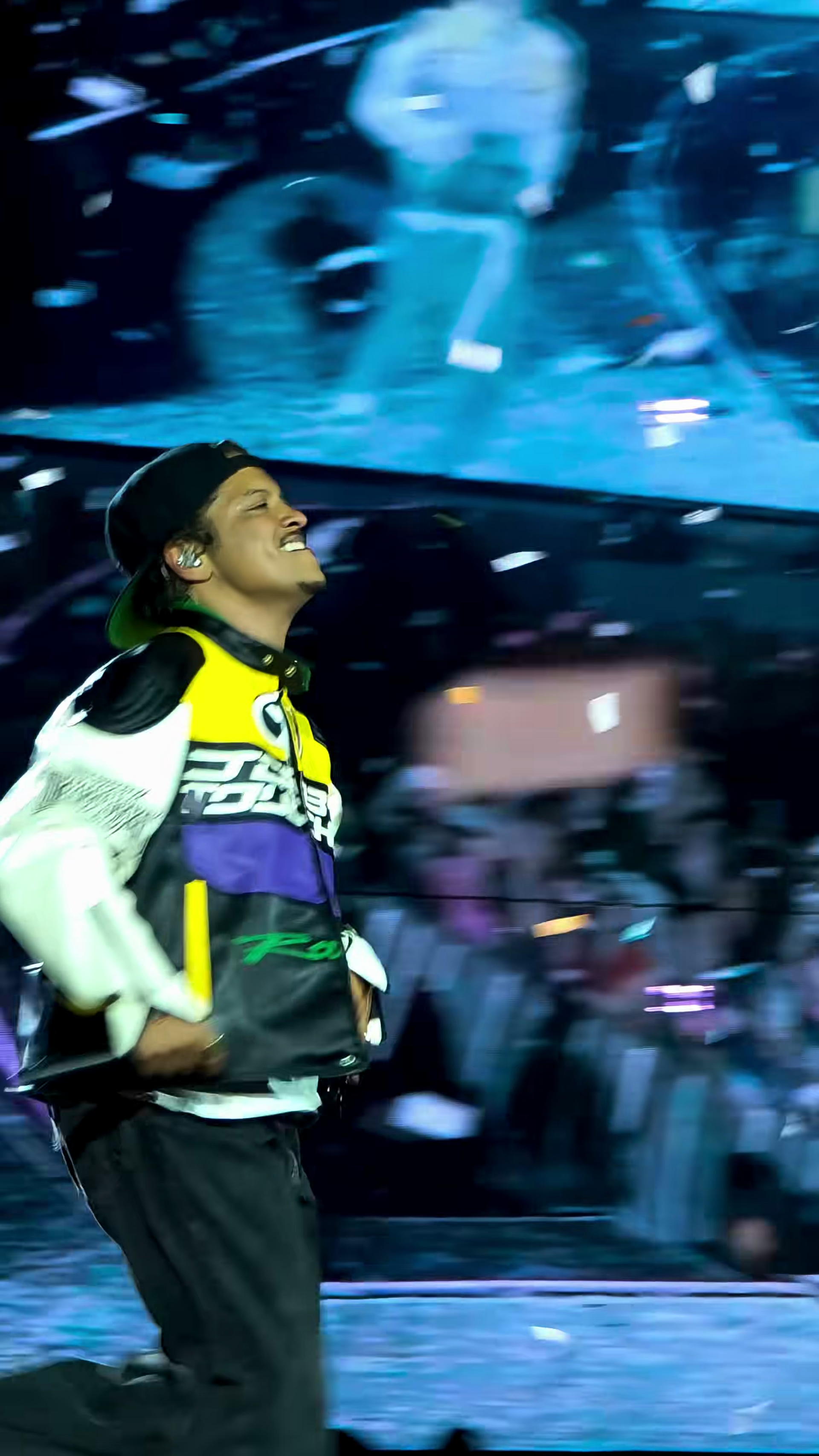 Bruno Mars laughs with his head tipped back on stage, in a backwards cap and a colour-blocked racing jacket, a video screen glowing behind him.