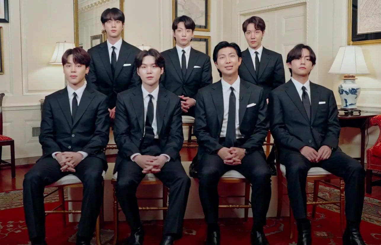 The seven members of BTS pose together in matching black suits and ties, seated and standing in a formal parlor.
