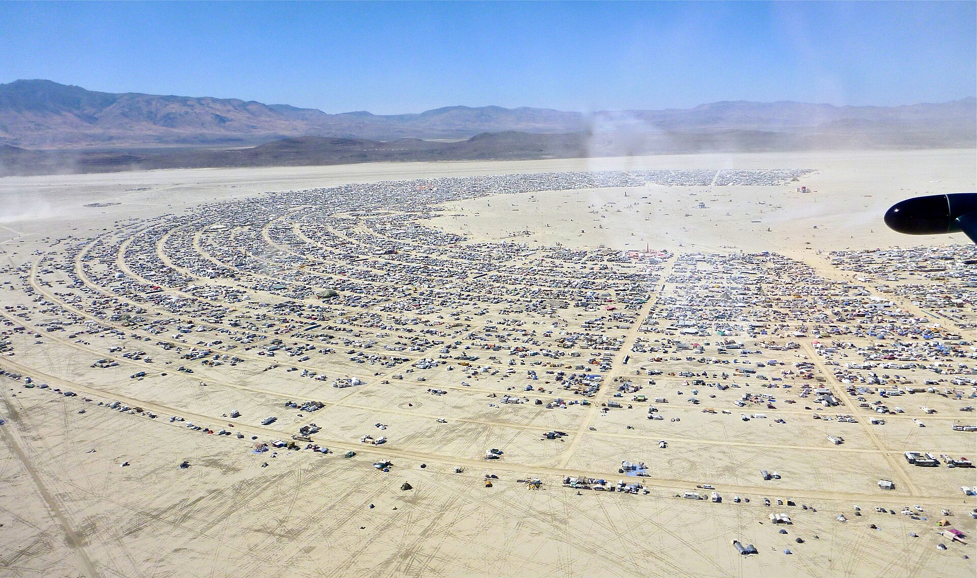 An aerial view of Black Rock City - thousands of tents, trailers and vehicles laid out in concentric arcs across a flat desert playa, mountains on the horizon.
