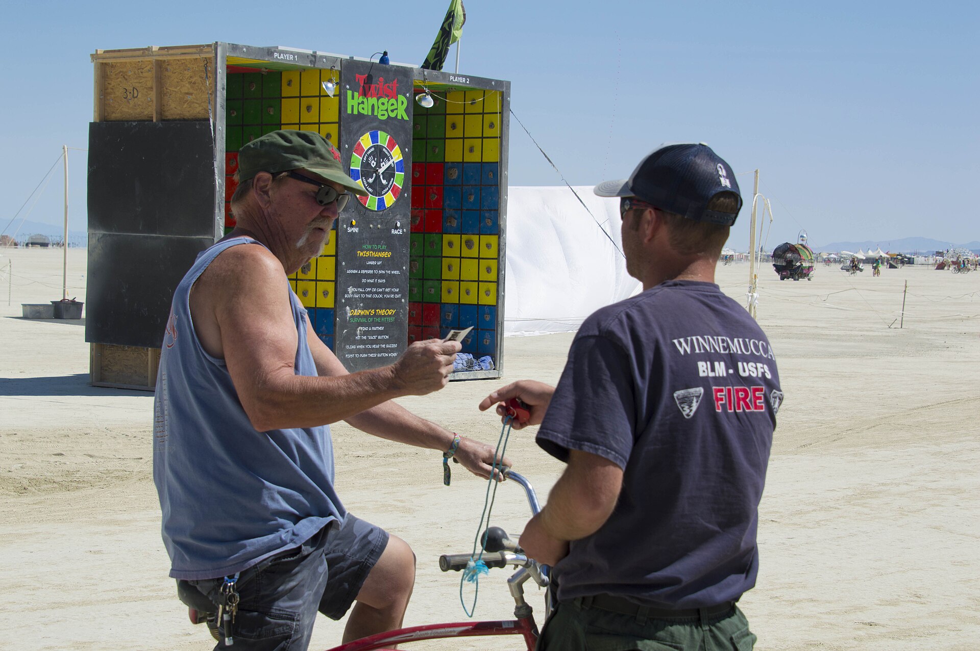 A man on a bicycle talks with another man in a BLM and Forest Service fire crew shirt on the open playa, beside a homemade carnival game booth.