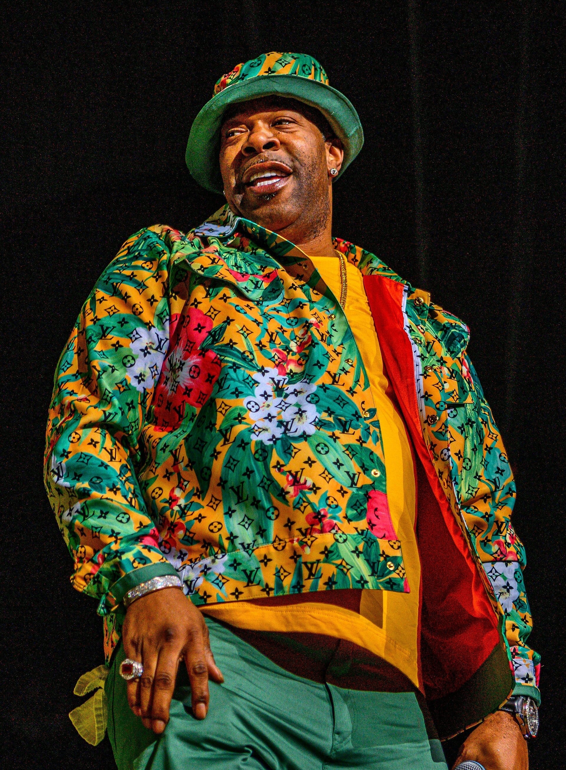 Busta Rhymes performs on stage, smiling and looking off to the side while holding a microphone, wearing a green bucket hat and a colorful floral jacket over a yellow shirt.