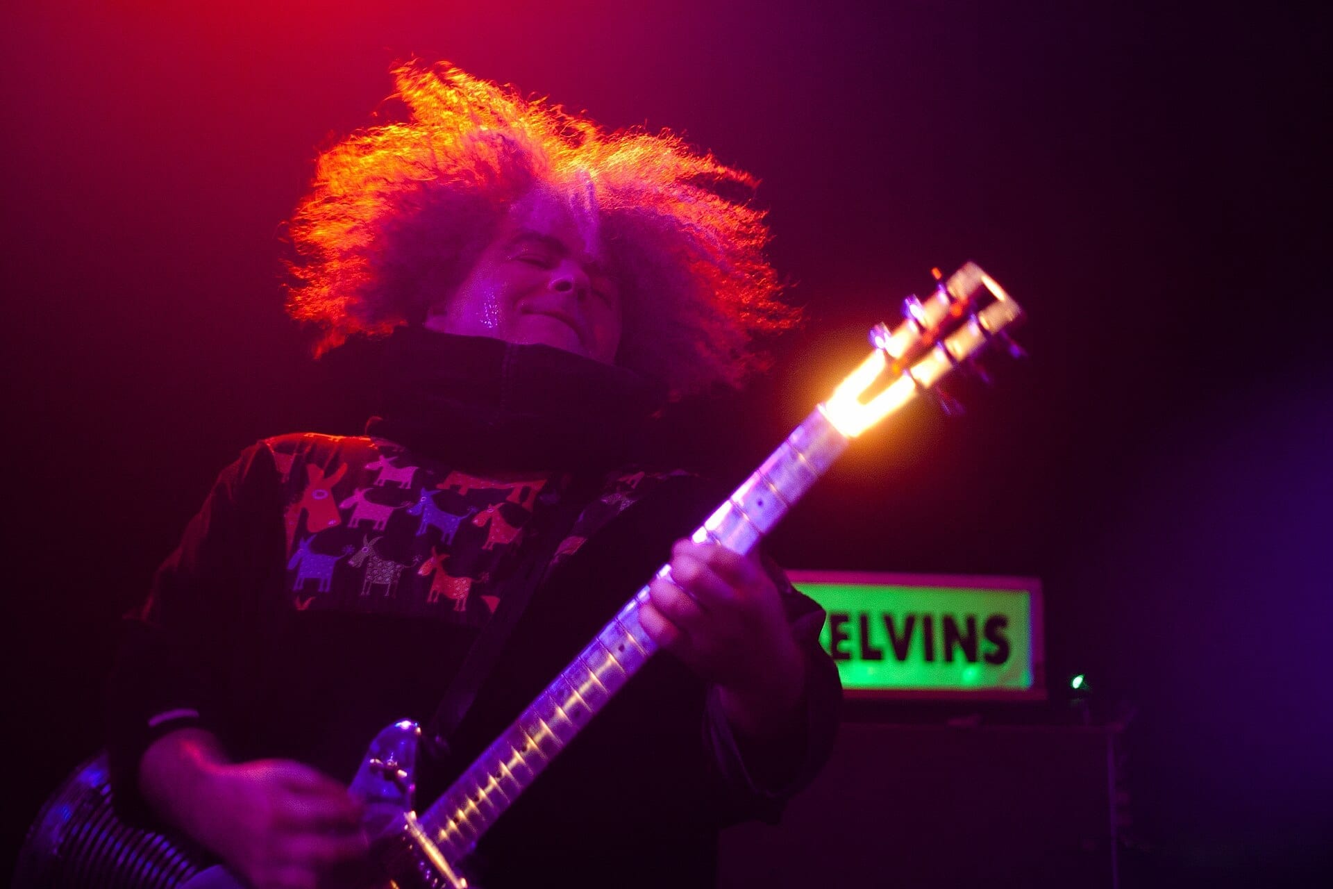 Buzz Osborne plays guitar with his head thrown back and hair flying, lit in red and purple, with a glowing green MELVINS sign behind him.