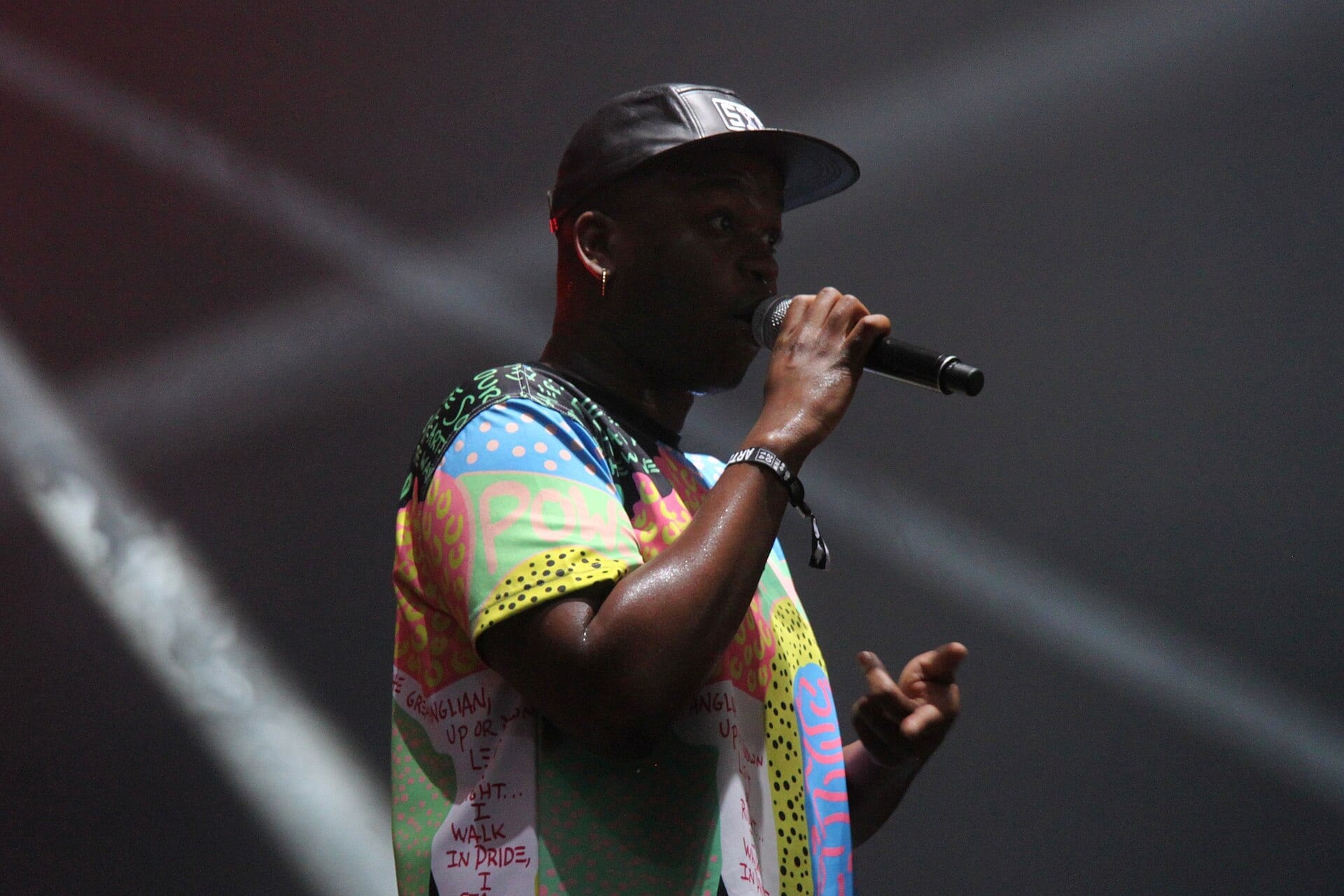 Cakes da Killa raps into a handheld microphone in a brightly patterned shirt and a black cap.