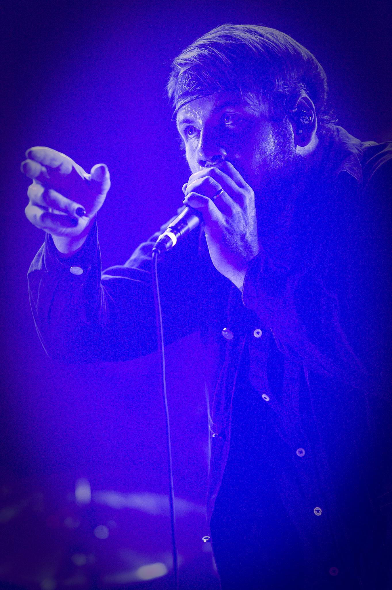 Beartooth's singer cups a handheld microphone and reaches out with his free hand, in a bandana headband under deep blue light.