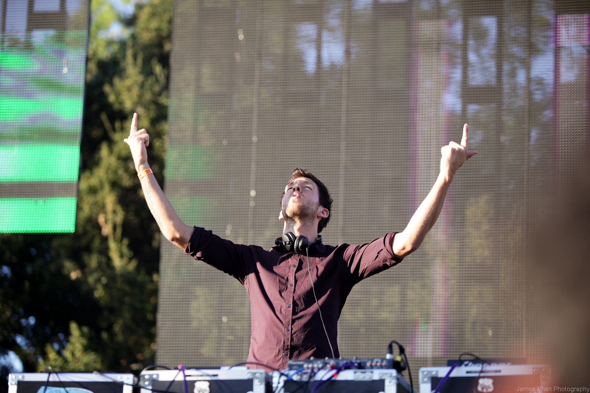 Calvin Harris stands at a DJ booth with both arms raised toward the sky and headphones hung around his neck, an outdoor stage screen behind him.