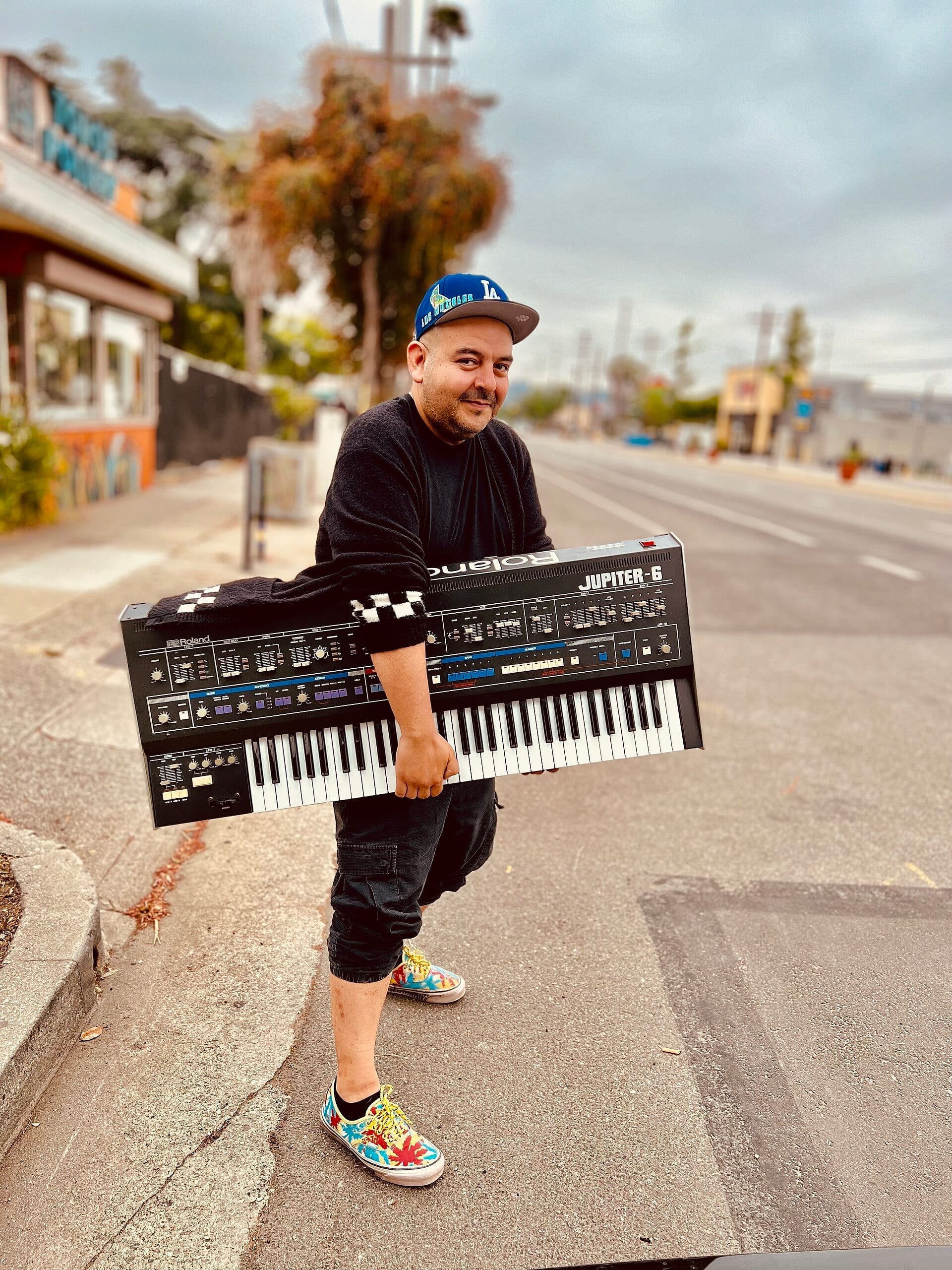 Camilo Lara, wearing an LA cap, carries a large Roland Jupiter-6 synthesizer keyboard on a Los Angeles street.