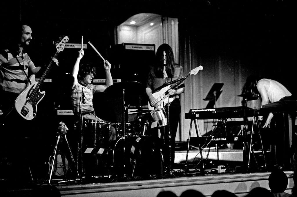 The band Can performs on stage with guitars, drums and a Farfisa keyboard, in a black and white photo.