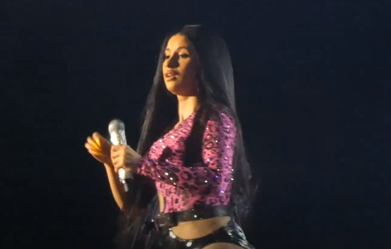 A rapper in a pink sequinned top with long dark hair holds a microphone at her side on a dark stage, looking off to one side