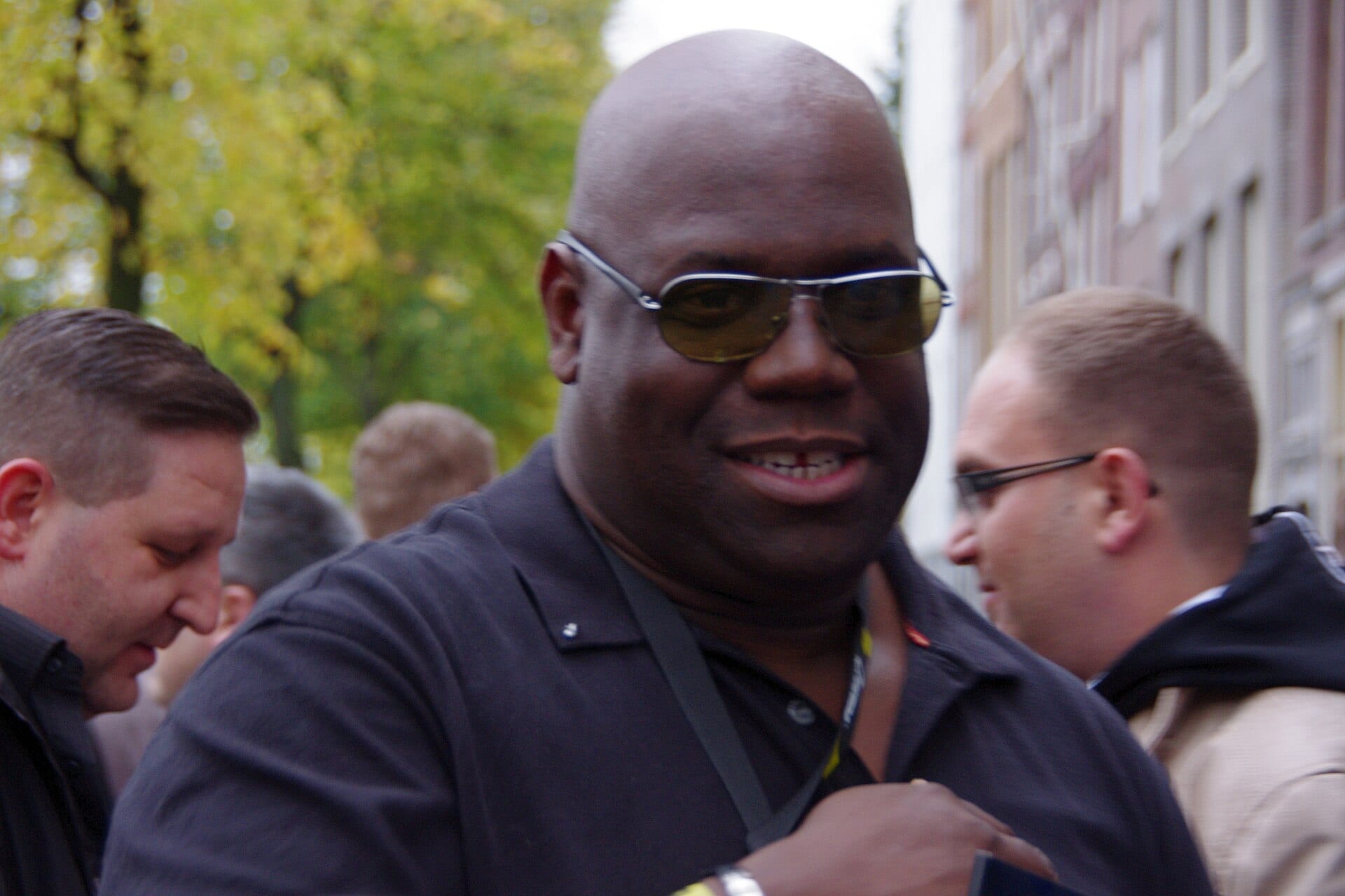 Carl Cox, wearing tinted aviator sunglasses and a dark shirt with a lanyard, smiles while walking through a crowd on a tree-lined street.
