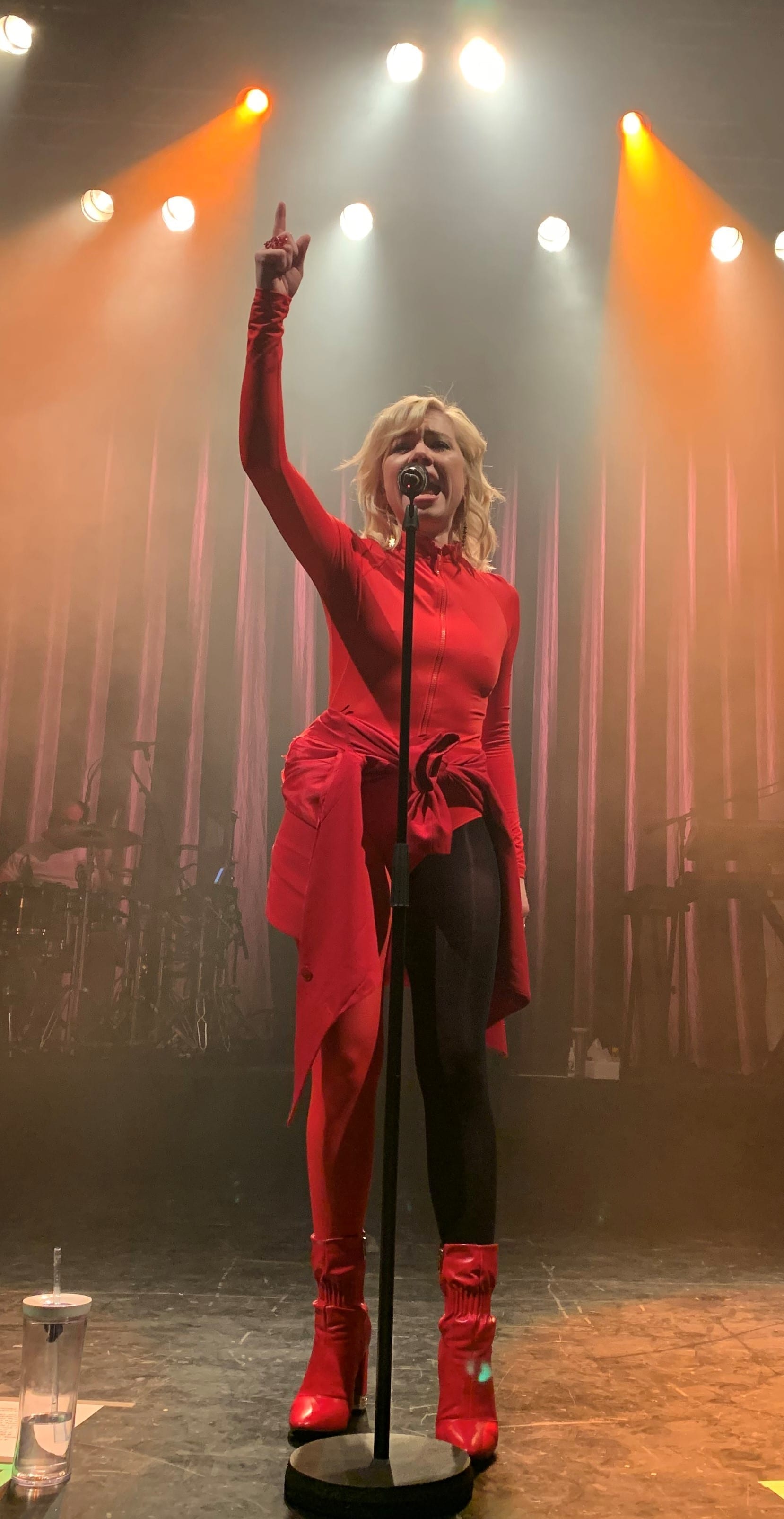 Carly Rae Jepsen, in an all-red outfit with red boots, sings into a microphone with one arm raised, backlit by warm stage lights and haze.