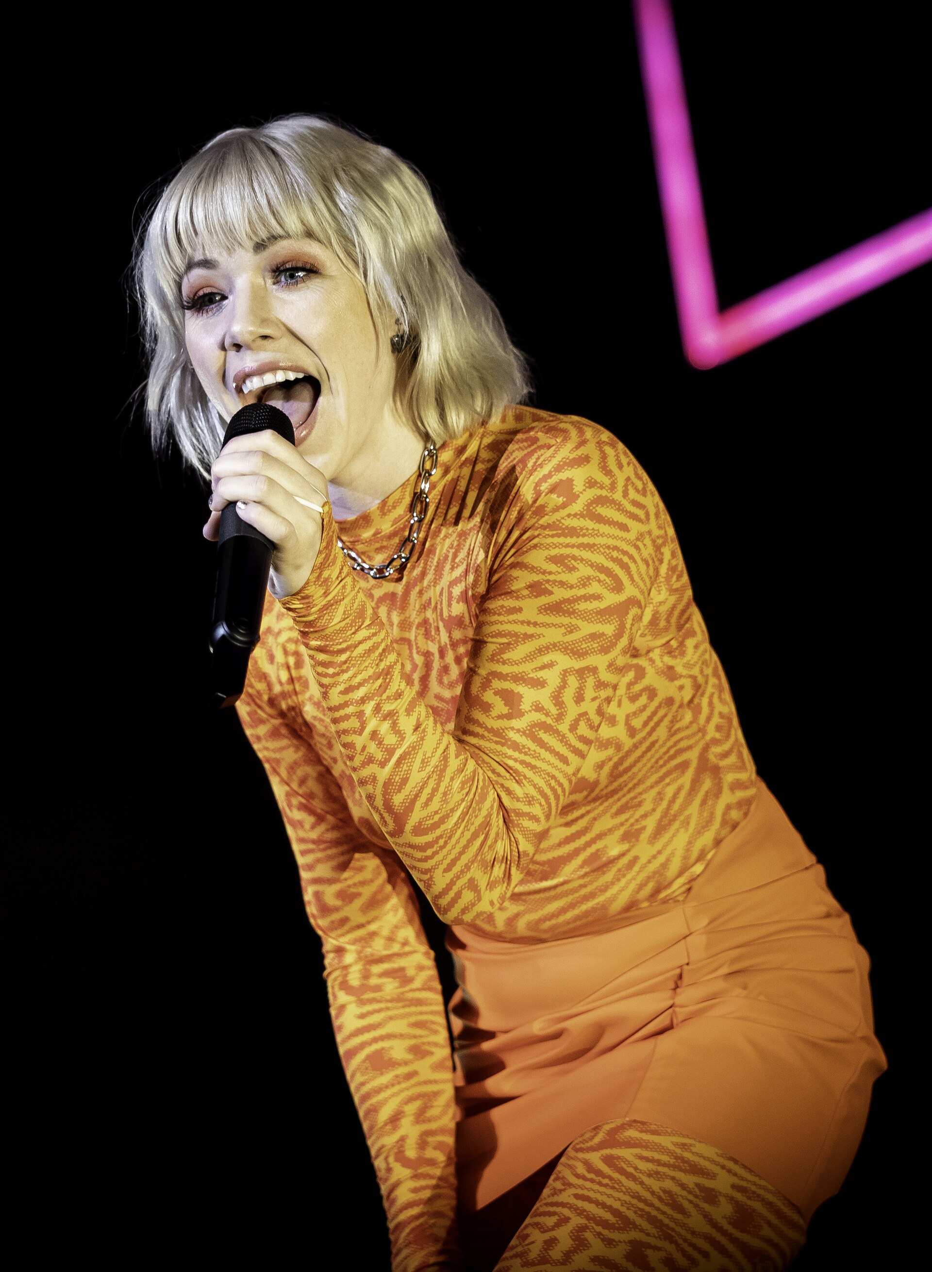 A blonde singer in an orange patterned outfit sings into a handheld microphone, a pink neon shape glowing behind her in the dark