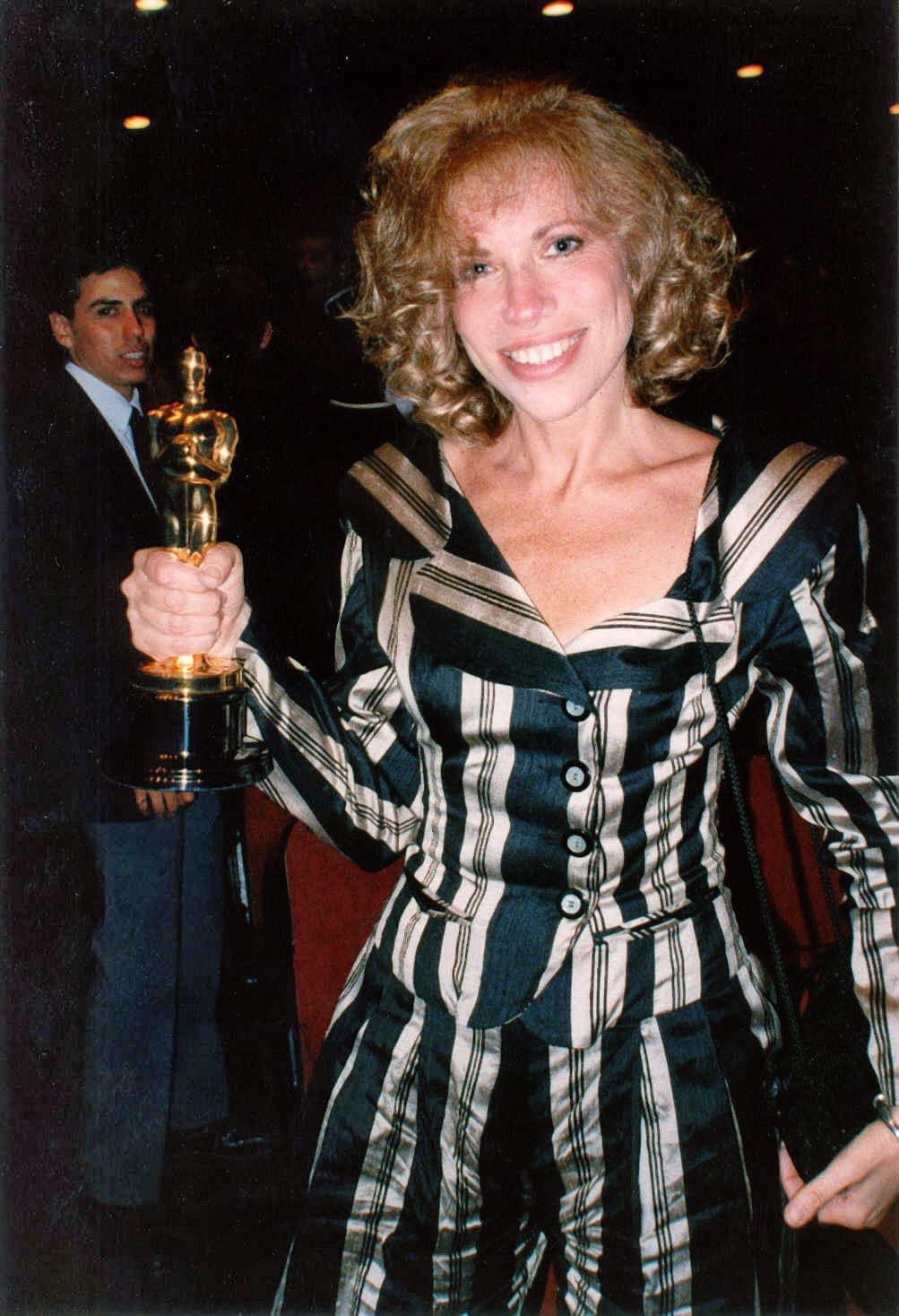 Carly Simon smiles while holding up an Academy Award trophy, wearing a striped black and white outfit.