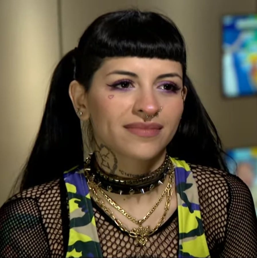 Cazzu, wearing a mesh top and layered gold necklaces, smiling during an interview.
