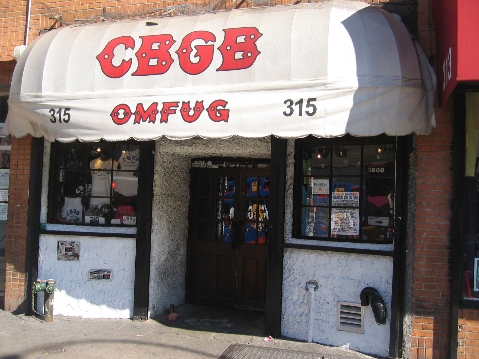 The front of CBGB at 315 Bowery, its white awning lettered CBGB and OMFUG in red, with merchandise in the two windows either side of the doorway.