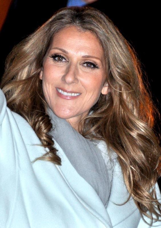 Celine Dion, with long wavy hair and a light gray coat, smiles at the camera in a close-up outdoor portrait.