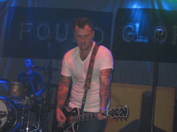 A guitarist in a white t-shirt with tattooed arms plays an electric guitar onstage, a drummer behind him and a New Found Glory banner across the back wall