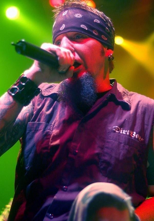 Chad Gray sings into a handheld microphone on a dimly lit stage, wearing a black bandana and a sleeveless button-up shirt that shows a full sleeve of tattoos on one arm.
