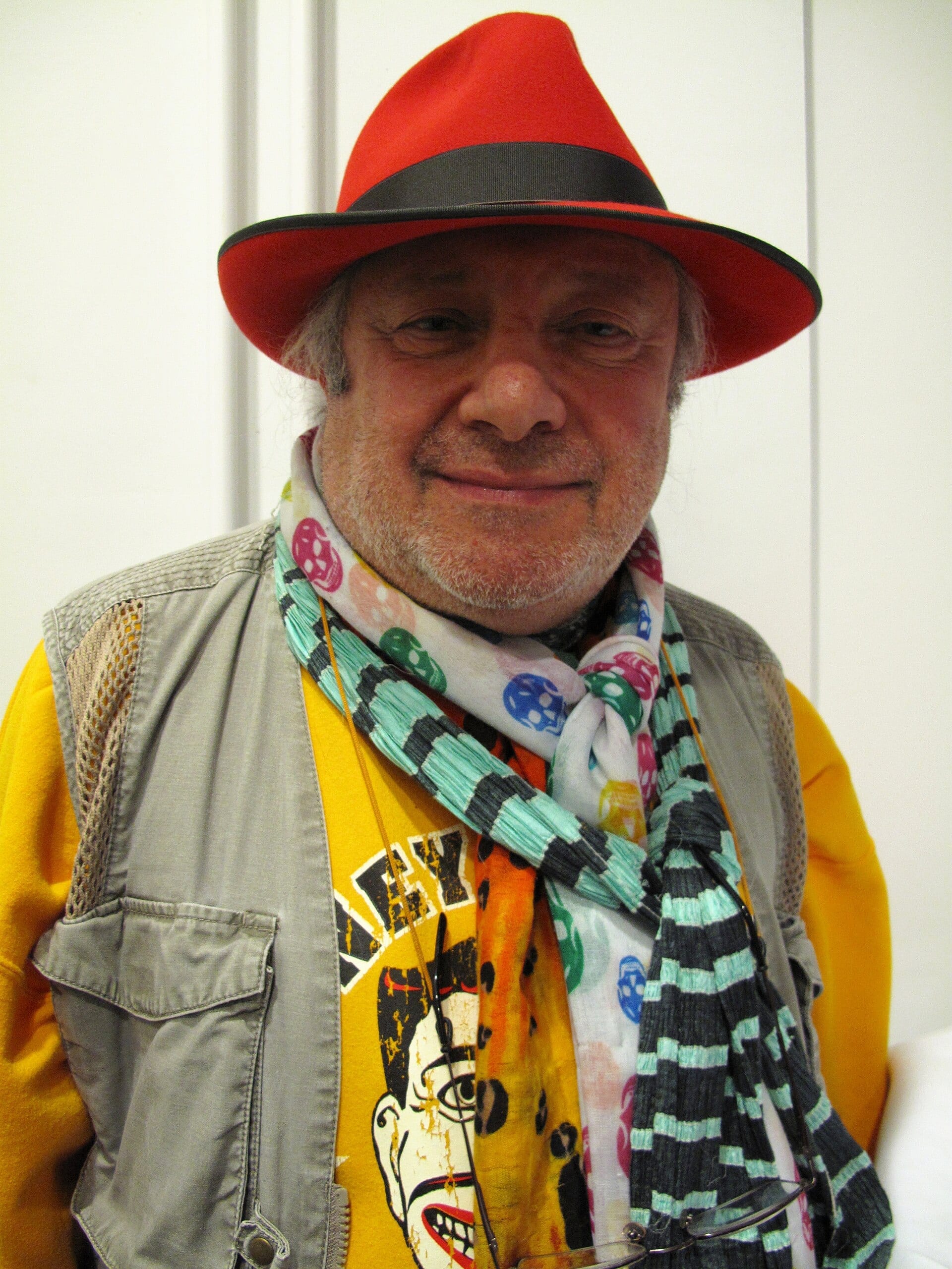 Charlemagne Palestine faces the camera in a red felt hat, a stack of patterned scarves around his neck over a yellow printed shirt and a grey utility vest.