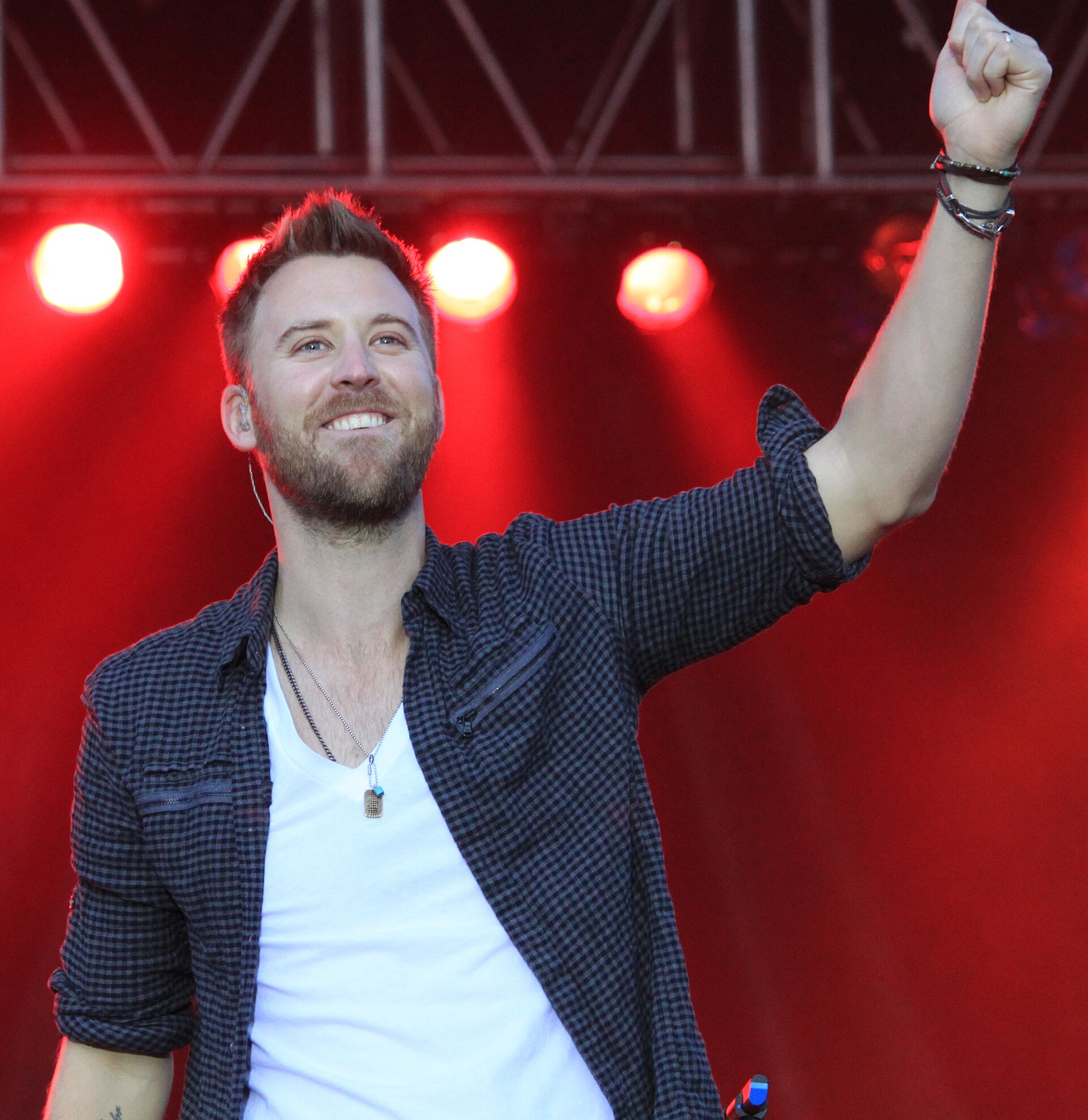 Charles Kelley grins and raises a fist on a red-lit stage, in an open checked shirt over a white T-shirt.
