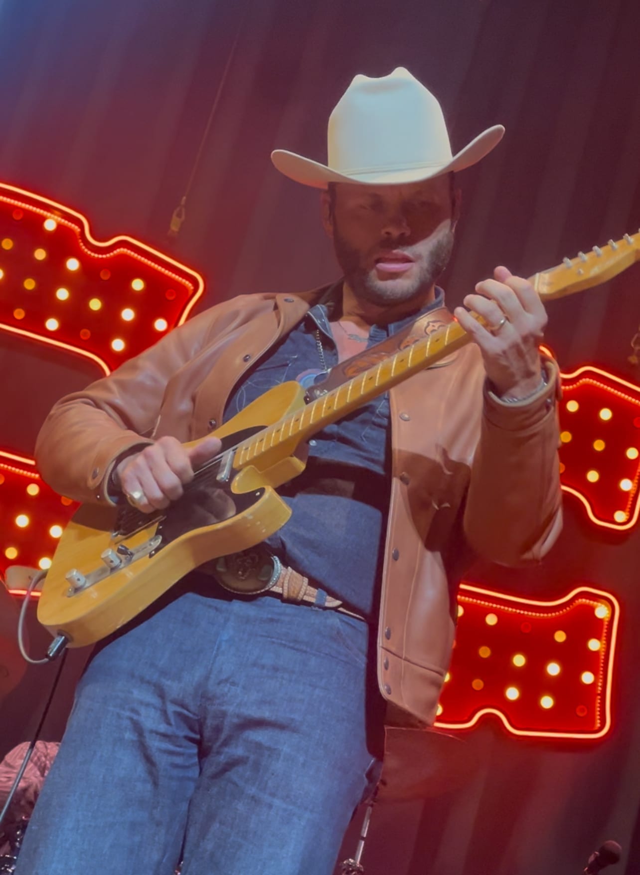 Charley Crockett plays a blond Telecaster in a white cowboy hat and tan leather jacket, red bulb-lit shapes glowing on the curtain behind him.