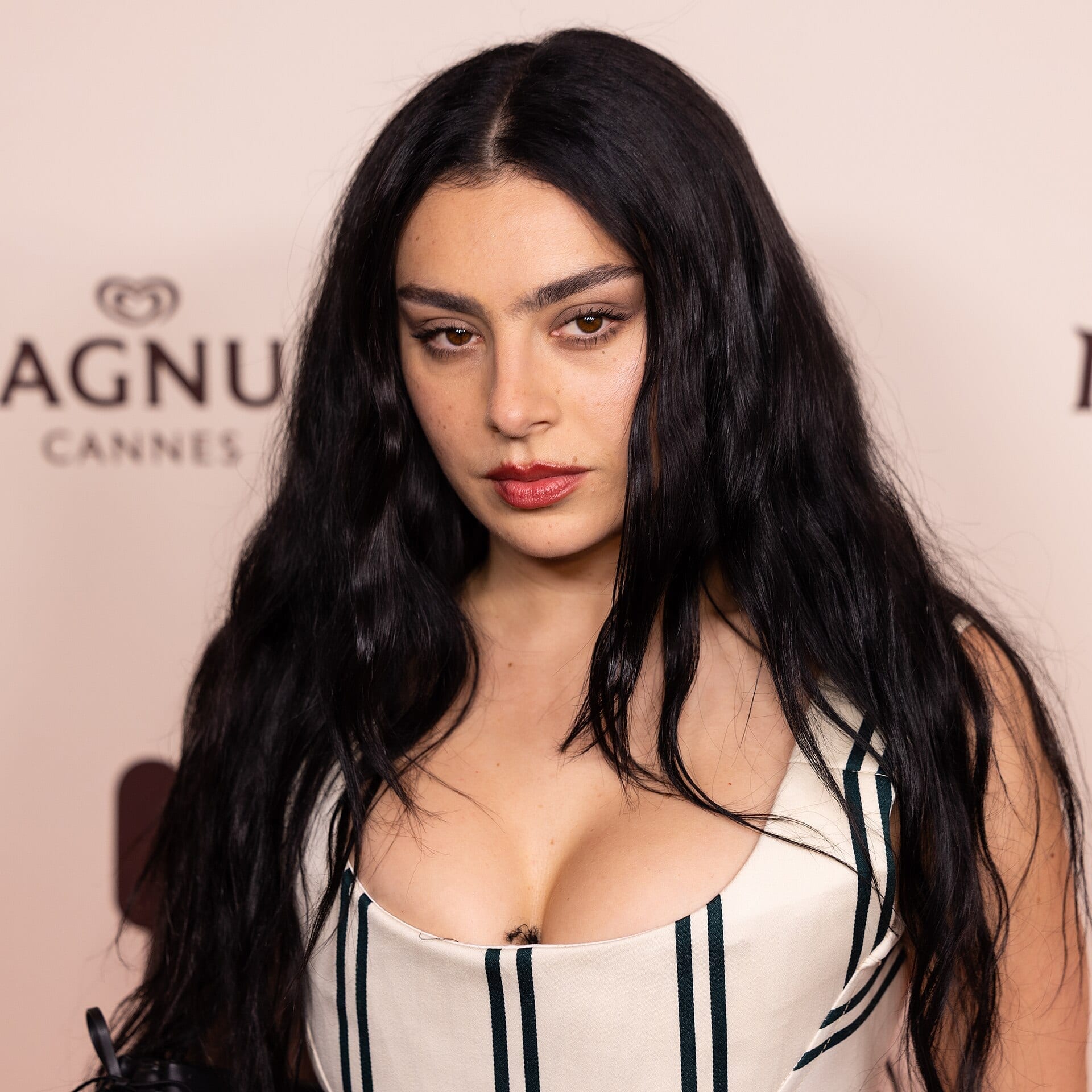 Charli XCX looks into the camera with dark wavy hair and red lipstick, wearing a white pinstriped dress in front of a step-and-repeat backdrop.