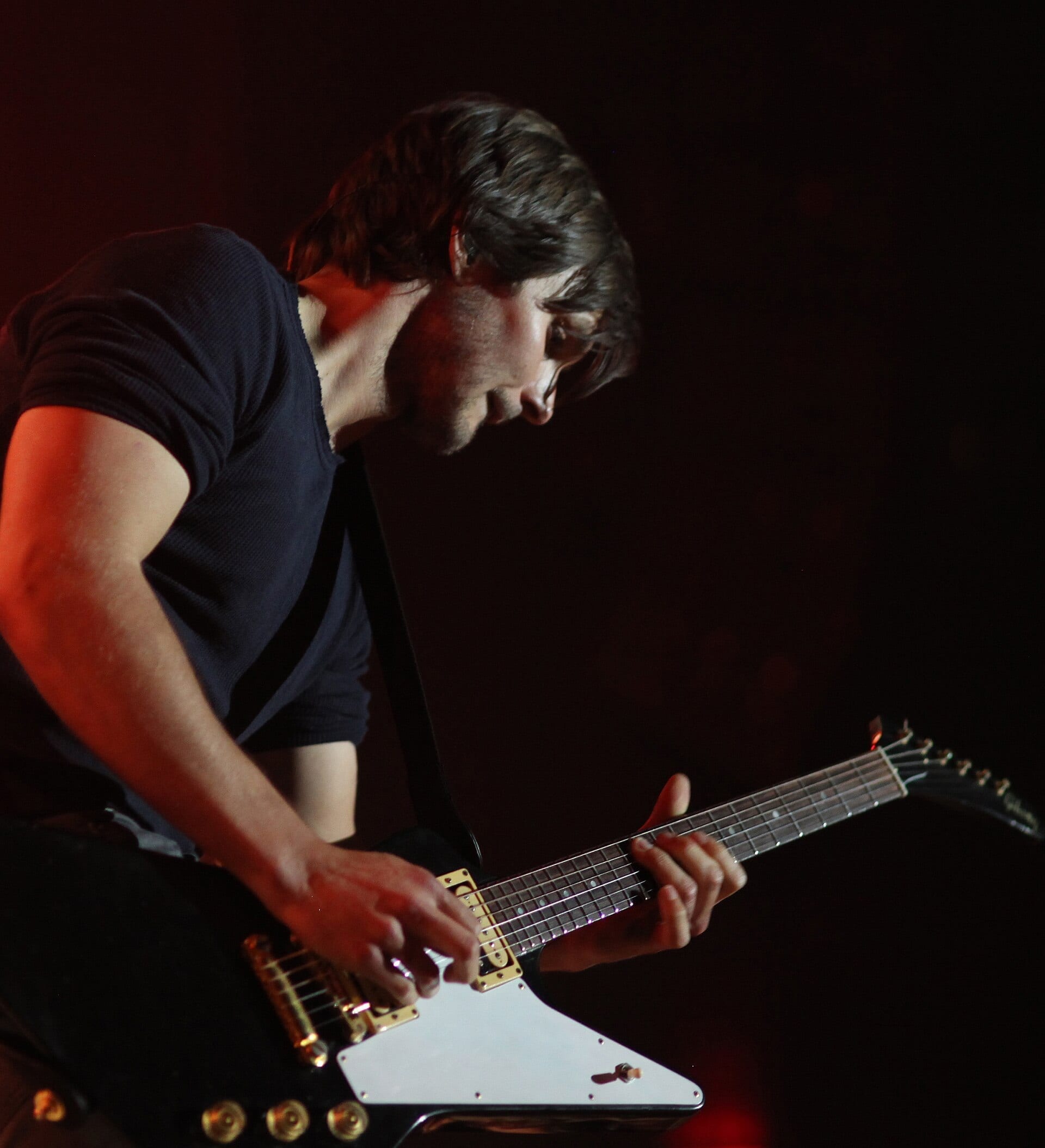 Charlie Worsham bends over a gold-hardware electric guitar under red stage light, his long hair falling forward as he picks the strings.