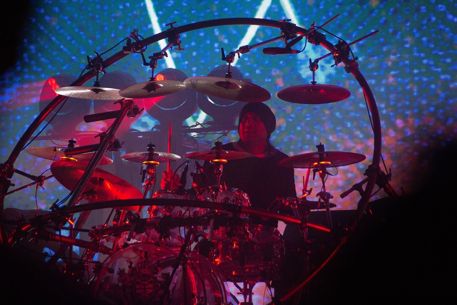 A drummer in a dark beanie plays a large ringed drum kit onstage, silhouetted red against a blue-lit LED backdrop.