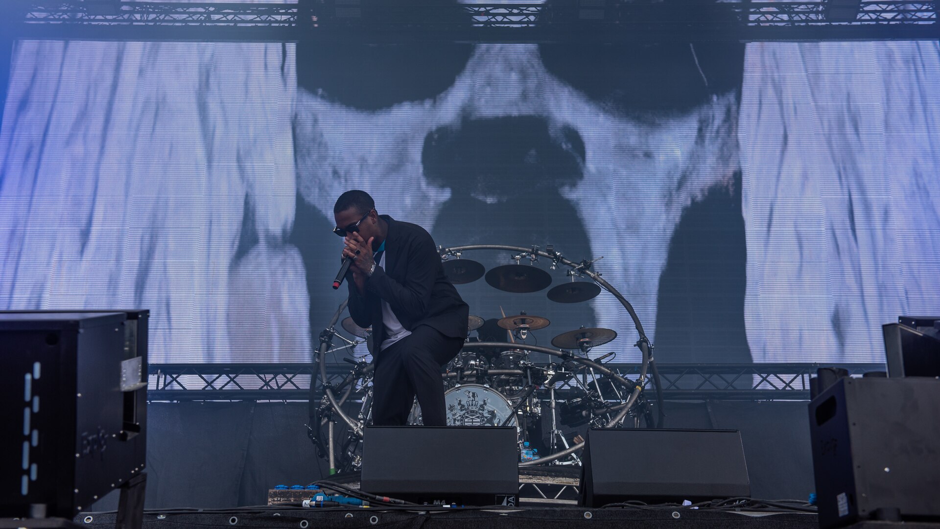 A vocalist in dark glasses and a jacket leans into a handheld microphone at the front of a Chase & Status stage, with the live drum kit lit behind him.
