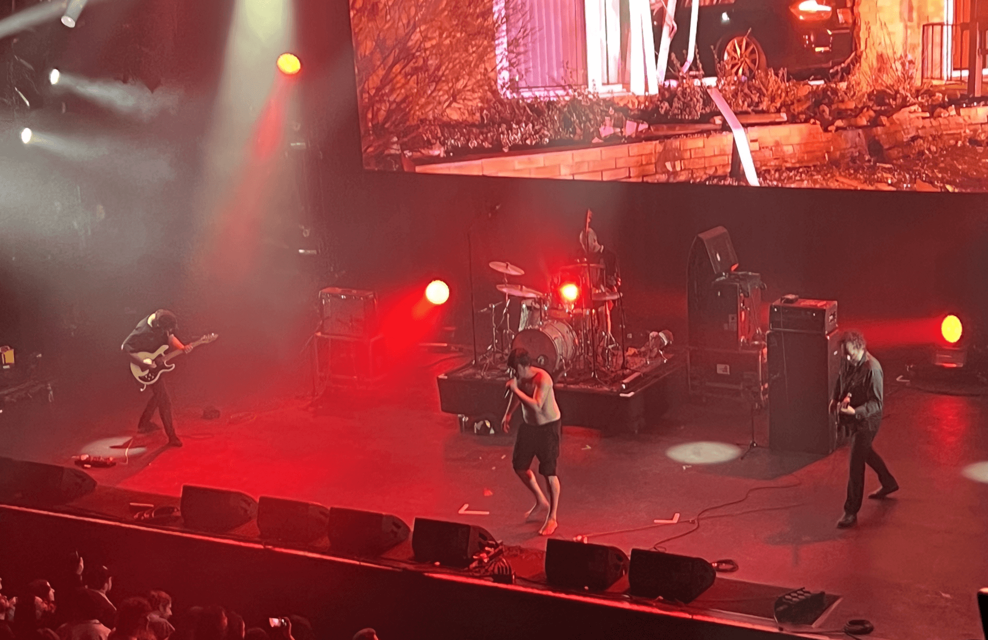 Chat Pile's shirtless singer crouches at a floor microphone under red stage lighting while two guitarists and a drummer play behind him, with a screen above the stage showing a suburban house scene.